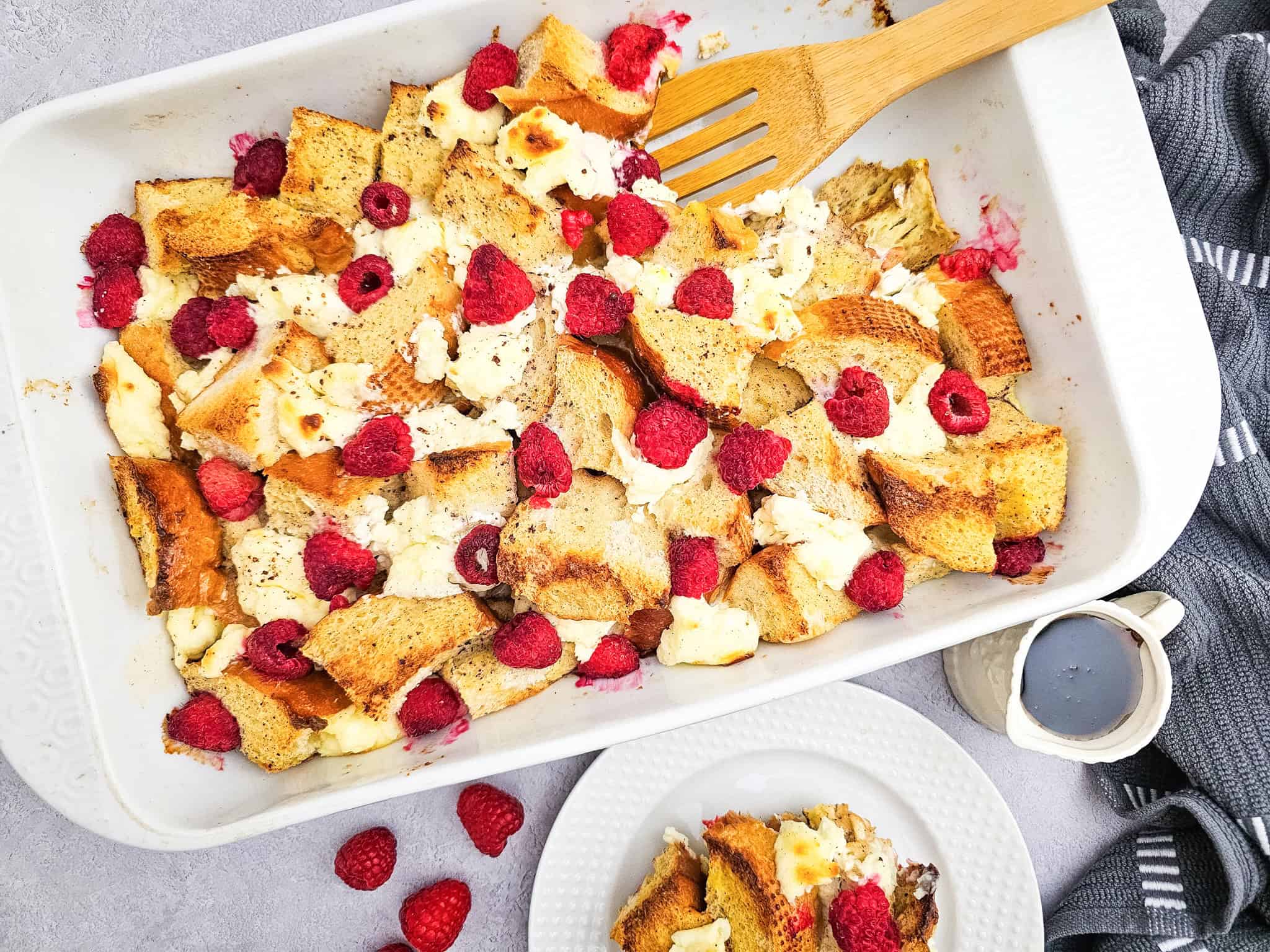 Top-down view of a full baking dish of raspberry French toast casserole. Golden chunks of bread, melty cream cheese, and juicy raspberries are ready to be served. A wooden spoon rests in the dish.