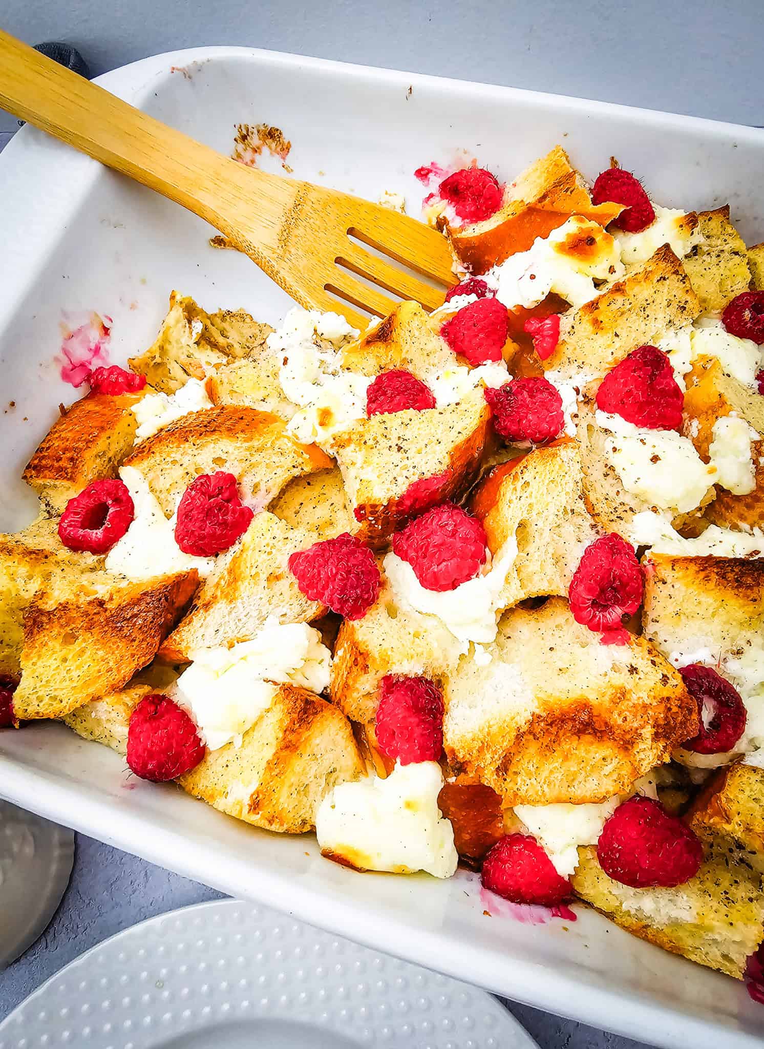 Side view of the casserole in the baking dish, showing the crispy, golden bread, rich cream cheese dollops, and vibrant raspberries throughout. A wooden spoon is tucked into the corner.