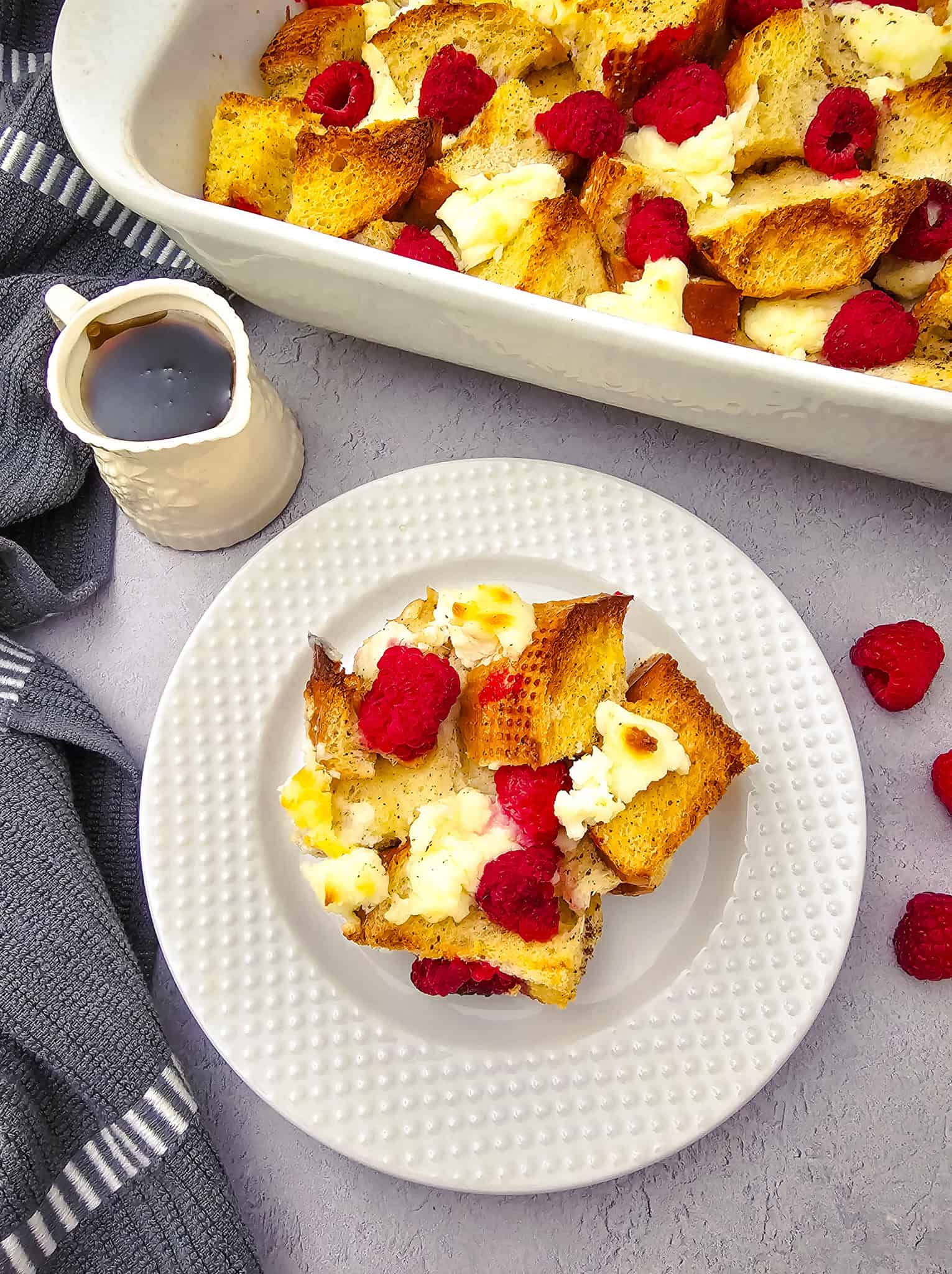 Full view of the casserole dish and a plated serving below, next to a syrup pitcher and scattered fresh raspberries. Warm, cozy colors give it a perfect brunch vibe.