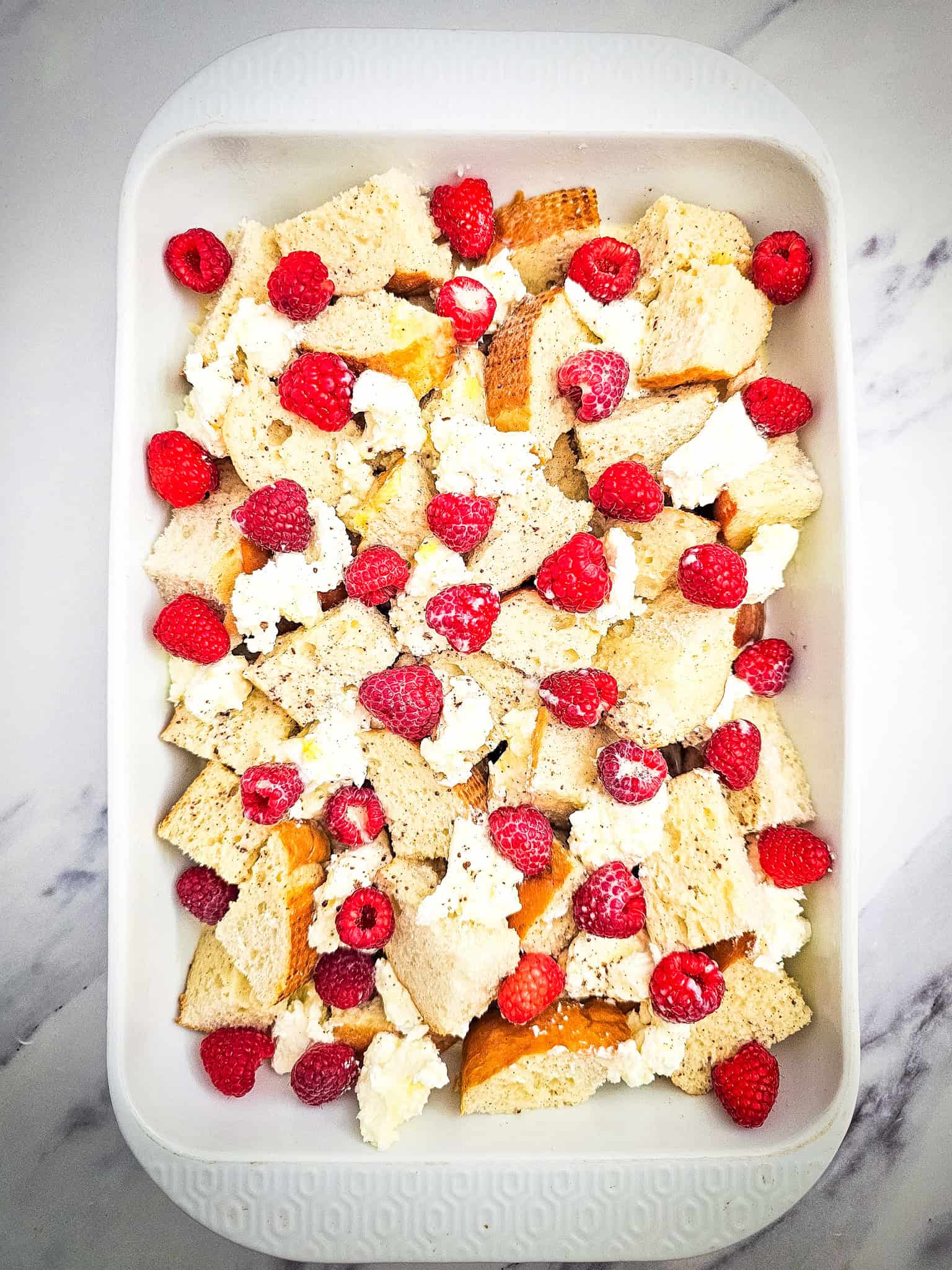 Top-down look at the unbaked French toast casserole, showing torn bread pieces topped with fresh raspberries and dollops of cream cheese in a white dish.