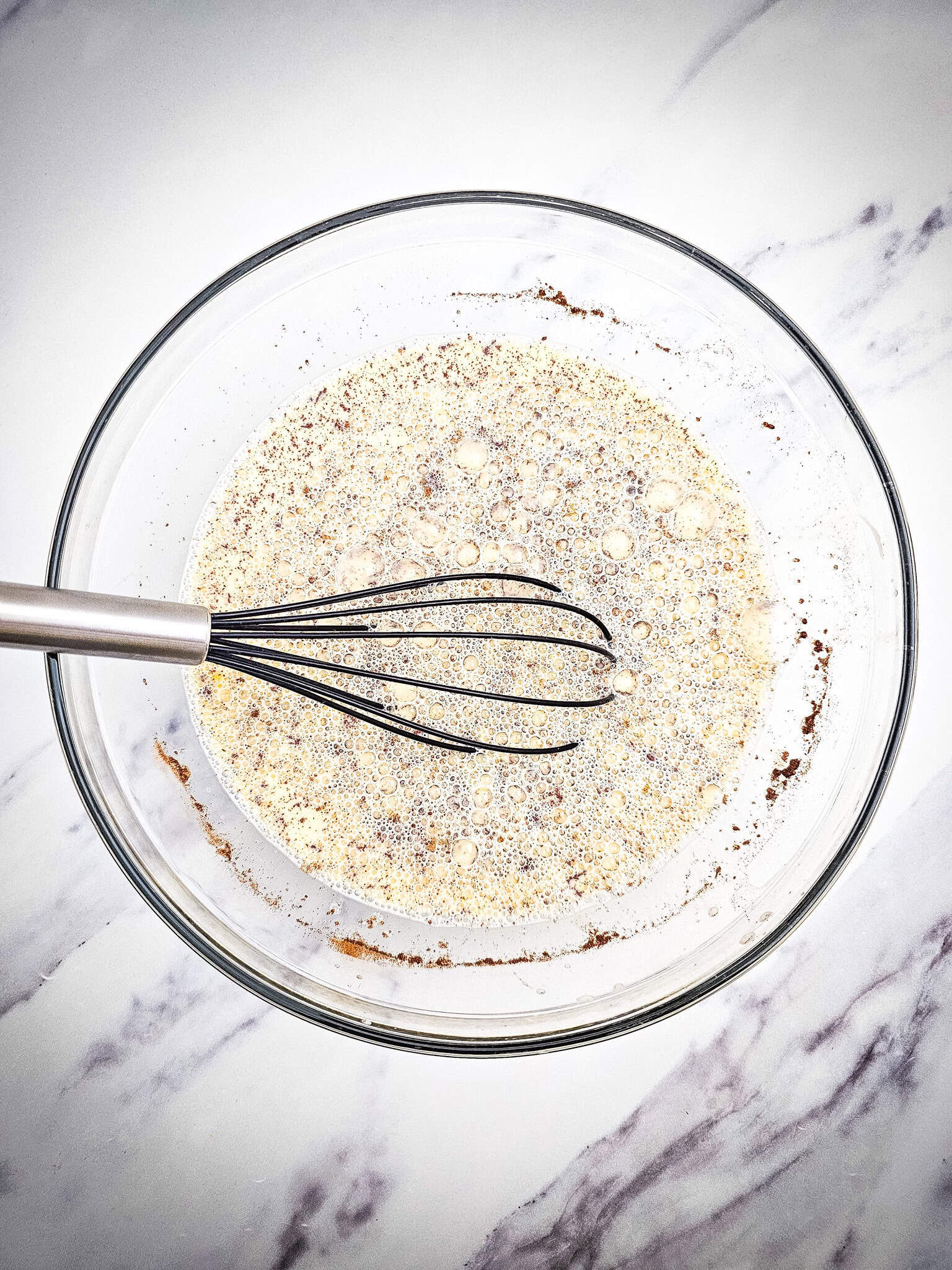 Glass mixing bowl with spiced egg and milk custard mixture, freshly whisked, ready to pour over the casserole ingredients. Cinnamon flecks are visible in the bubbly surface.