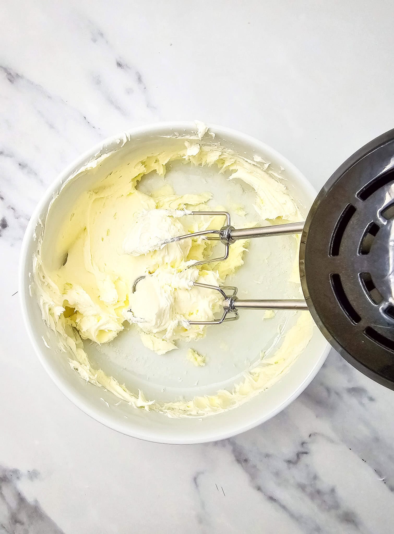 White mixing bowl with whipped cream cheese mixture and beaters resting on the side, ready to be spooned into the casserole.