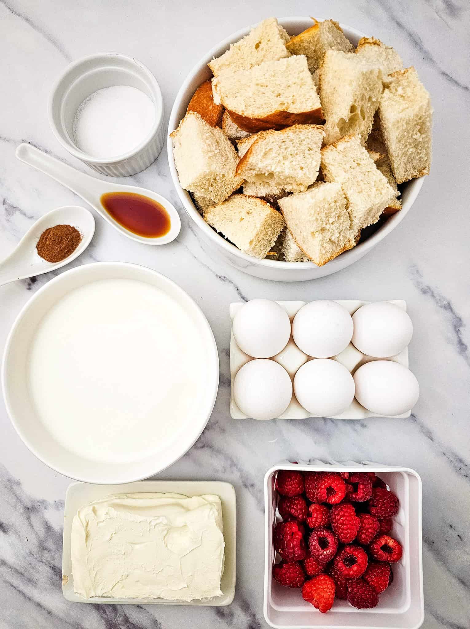 Overhead shot of all ingredients for raspberry French toast casserole: bread cubes, eggs, milk, cream cheese, raspberries, sugar, vanilla, and cinnamon, arranged on a marble surface.