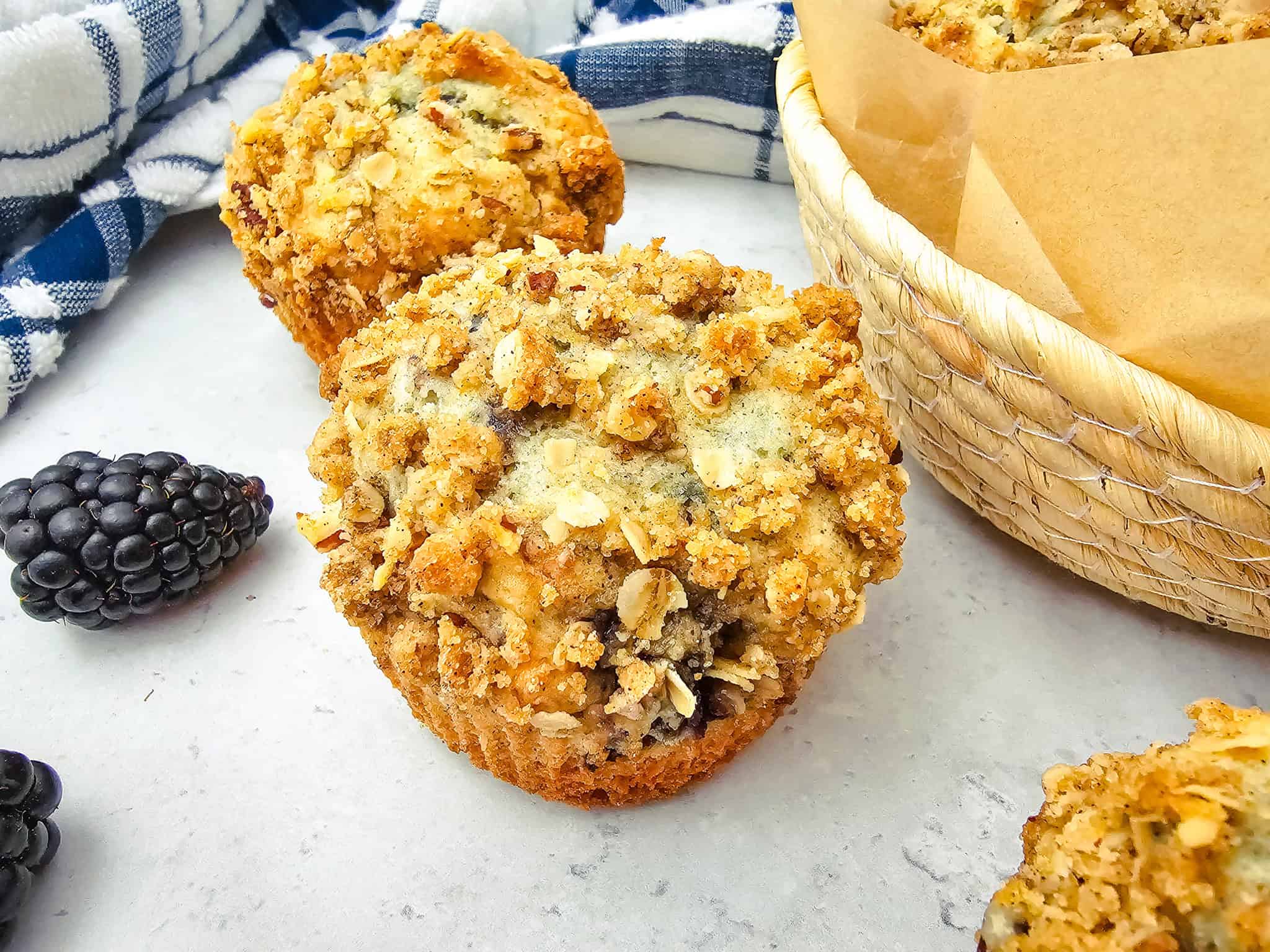 Close-up of a blackberry muffin with crumb topping, next to fresh blackberries and a basket of blackberry muffins—perfect inspiration for your next muffins recipe.