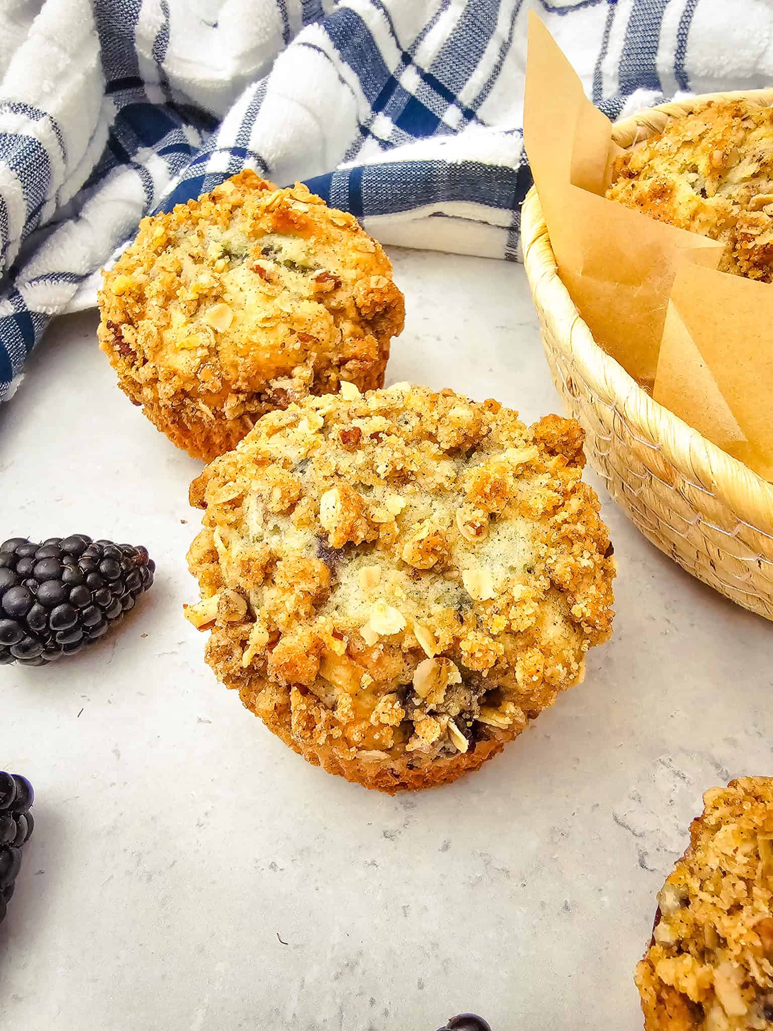 Two blackberry muffins with crumb topping sit on a countertop near fresh blackberries and a woven basket, perfect for anyone searching for the ultimate Blackberry Muffins recipe.