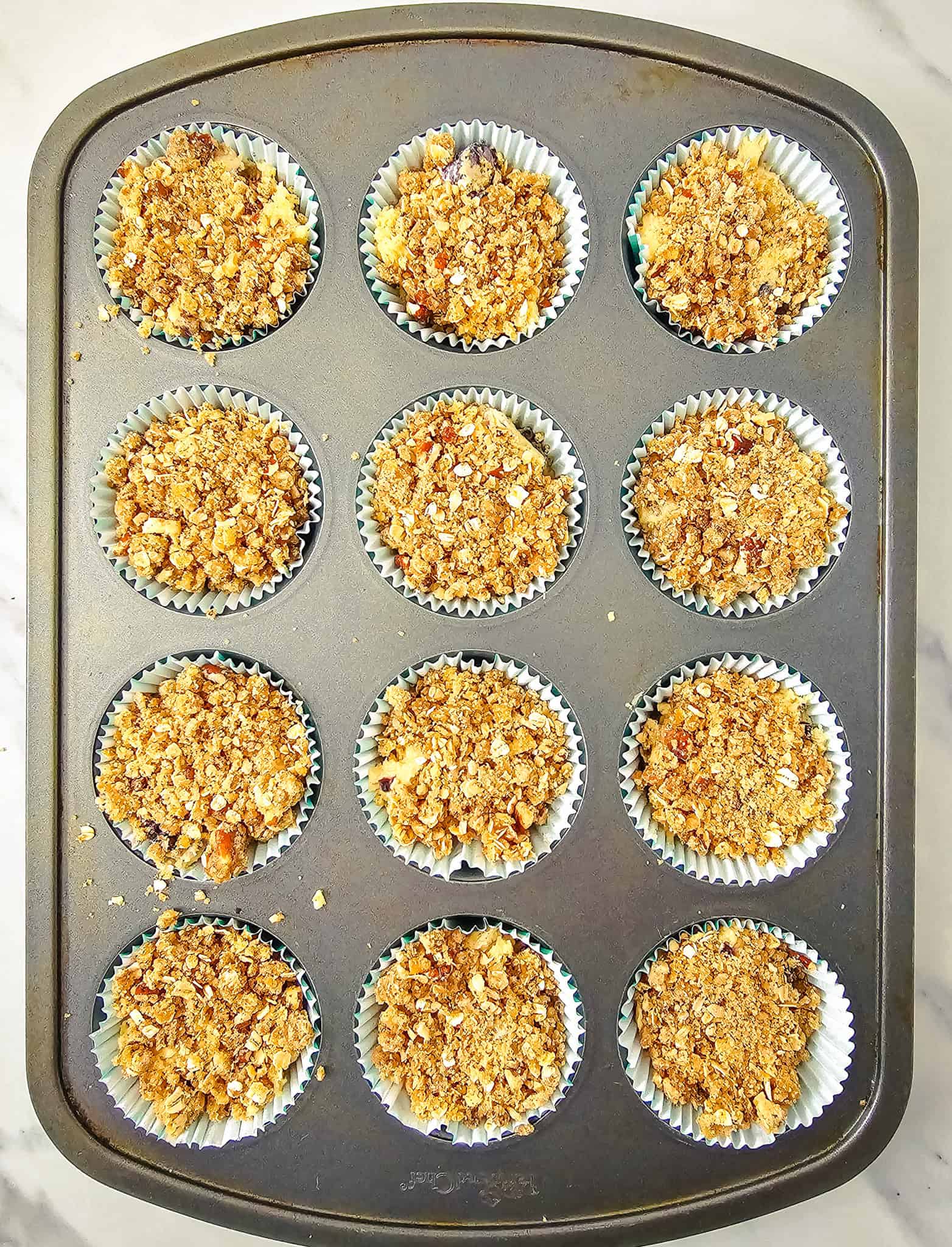 A muffin tin with 12 paper liners filled with oat and nut crumb-topped blackberry muffins batter, ready to bake—perfect for trying a new blackberry muffin recipe.