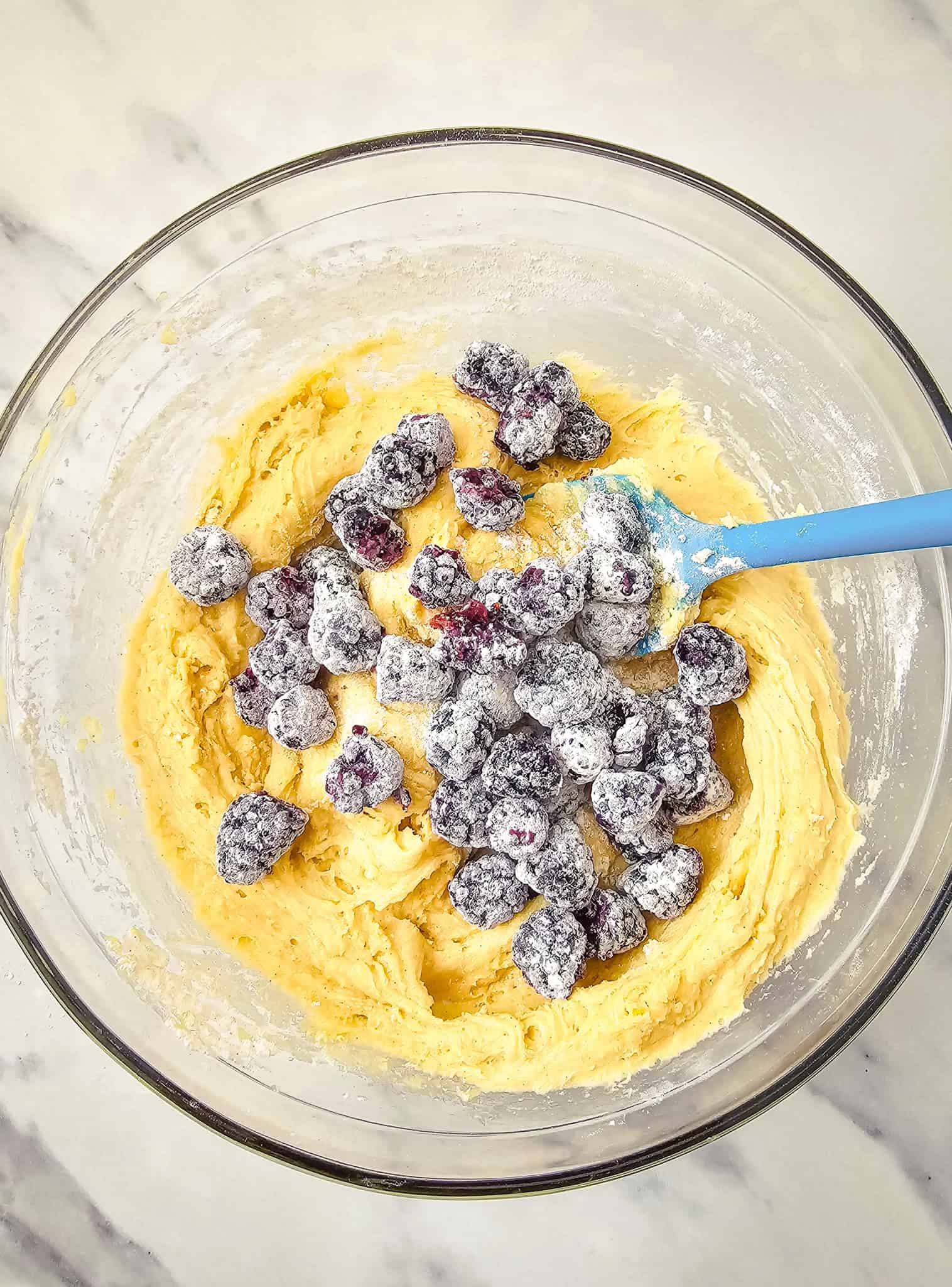 A bowl of cookie dough for a delicious Blackberry Muffins Recipe, with flour-dusted berries and a blue spatula on a marble countertop.