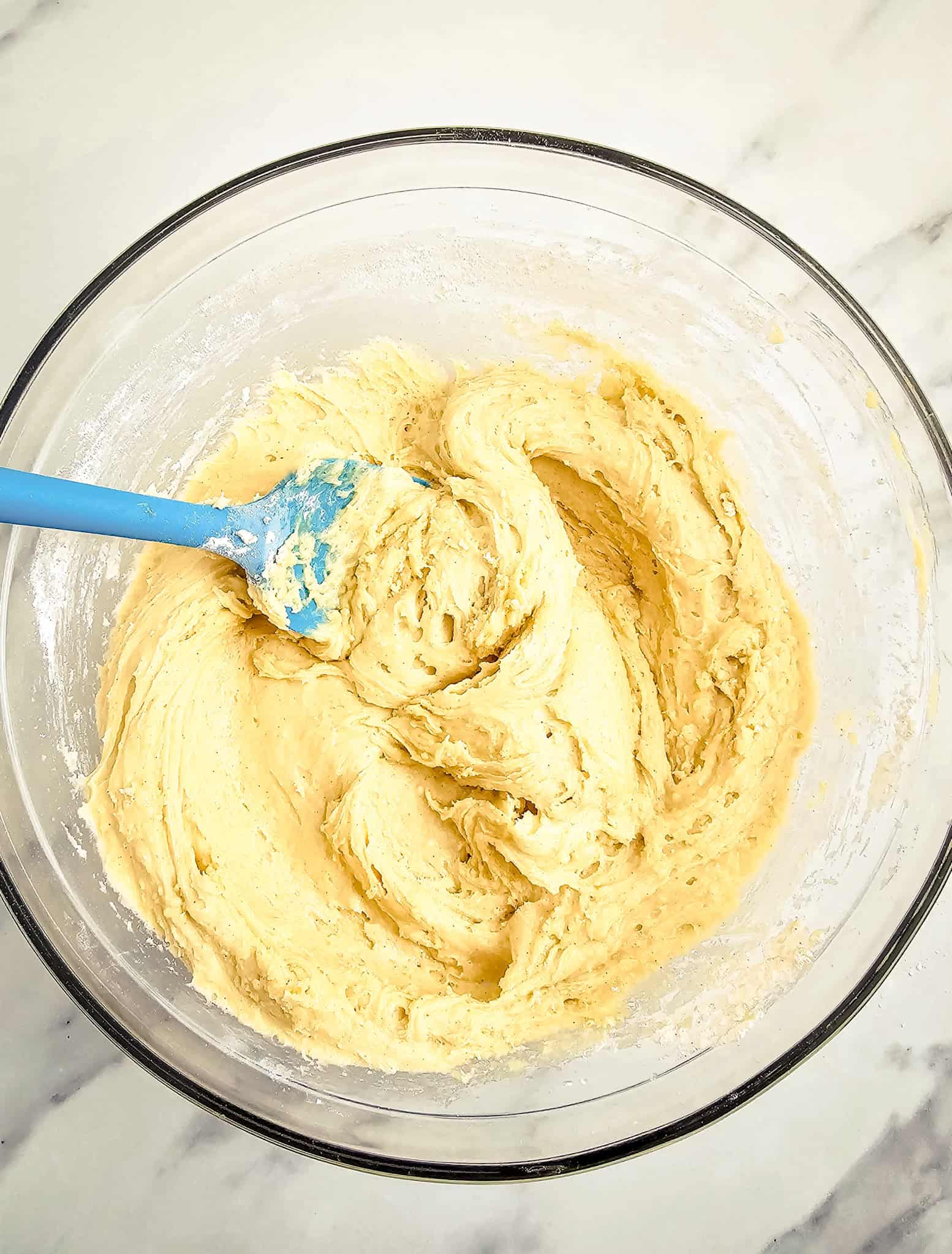 Thick yellow cake batter for a Blackberry Muffins Recipe being mixed with a blue spatula in a glass bowl on a marble surface.
