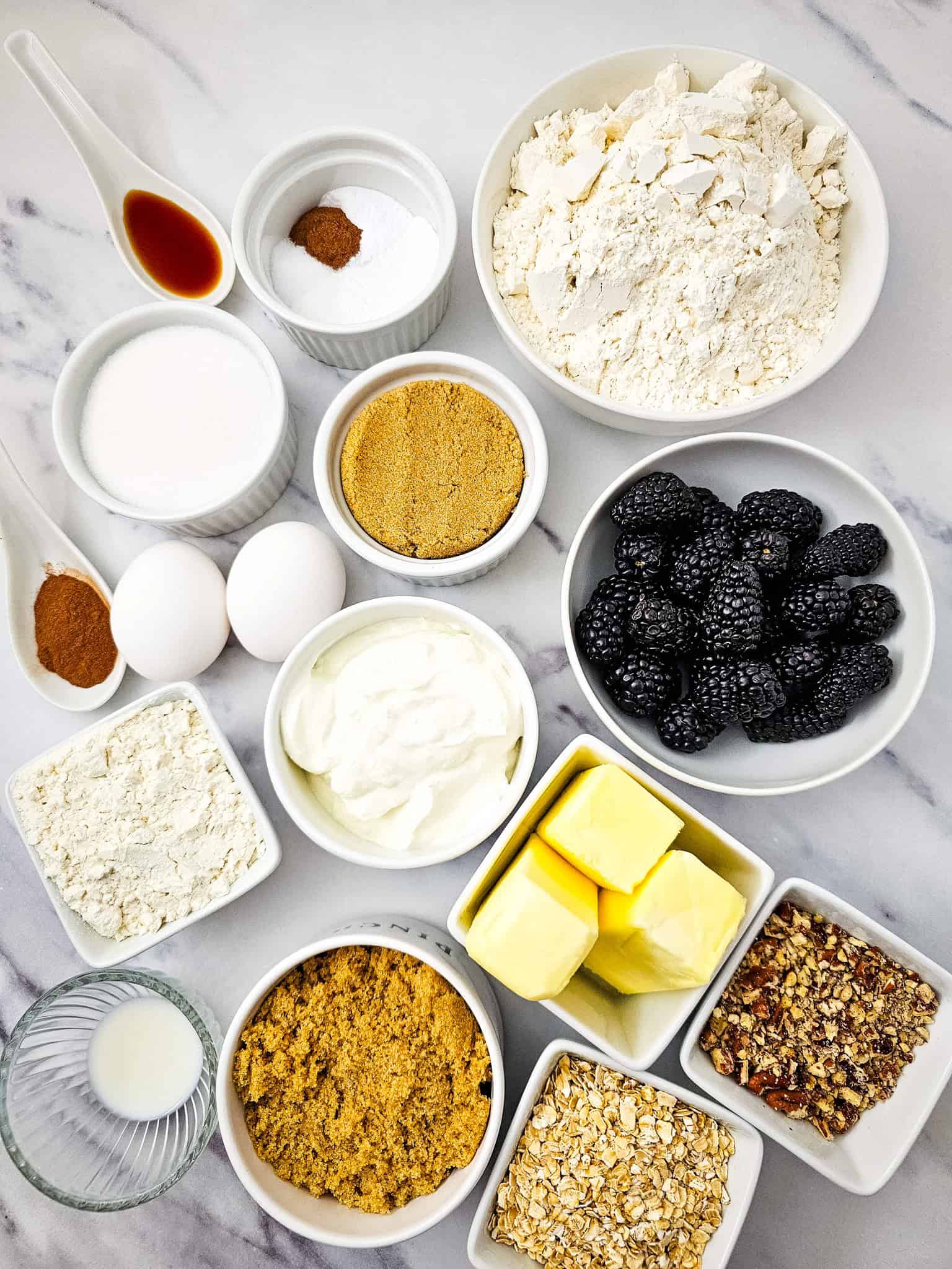 Baking ingredients in bowls on a marble surface, including flour, sugars, butter, eggs, blackberries, and spices