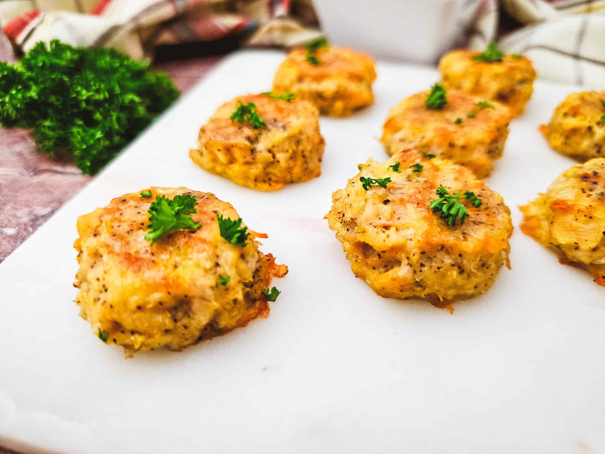 Baked chicken nuggets topped with parsley on a white cutting board with fresh herbs in the background.