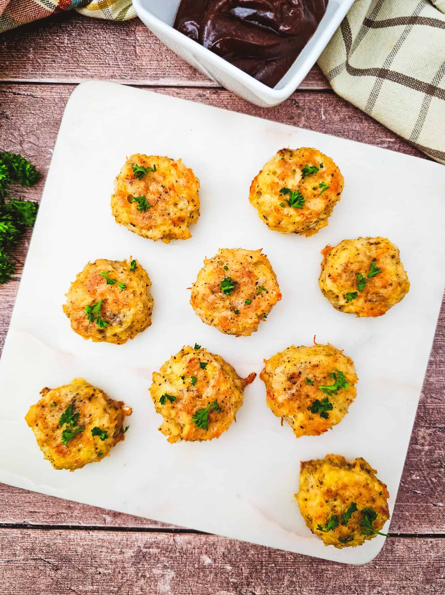 Nine baked low carb chicken nuggets, perfect as keto chicken nuggets, are arranged on a white board with a bowl of dark dipping sauce nearby.