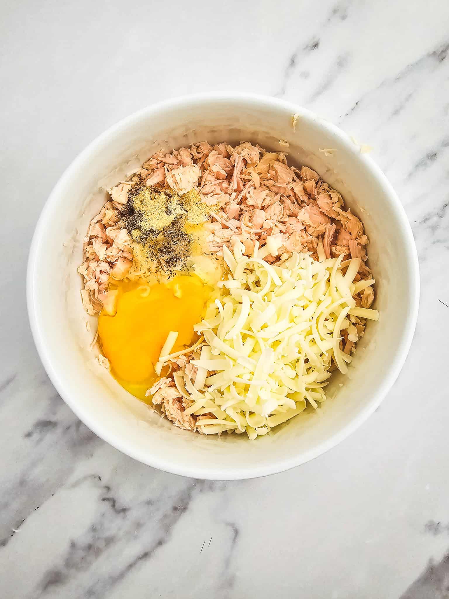 White bowl with shredded chicken, grated cheese, a raw egg, and seasonings on a marble surface