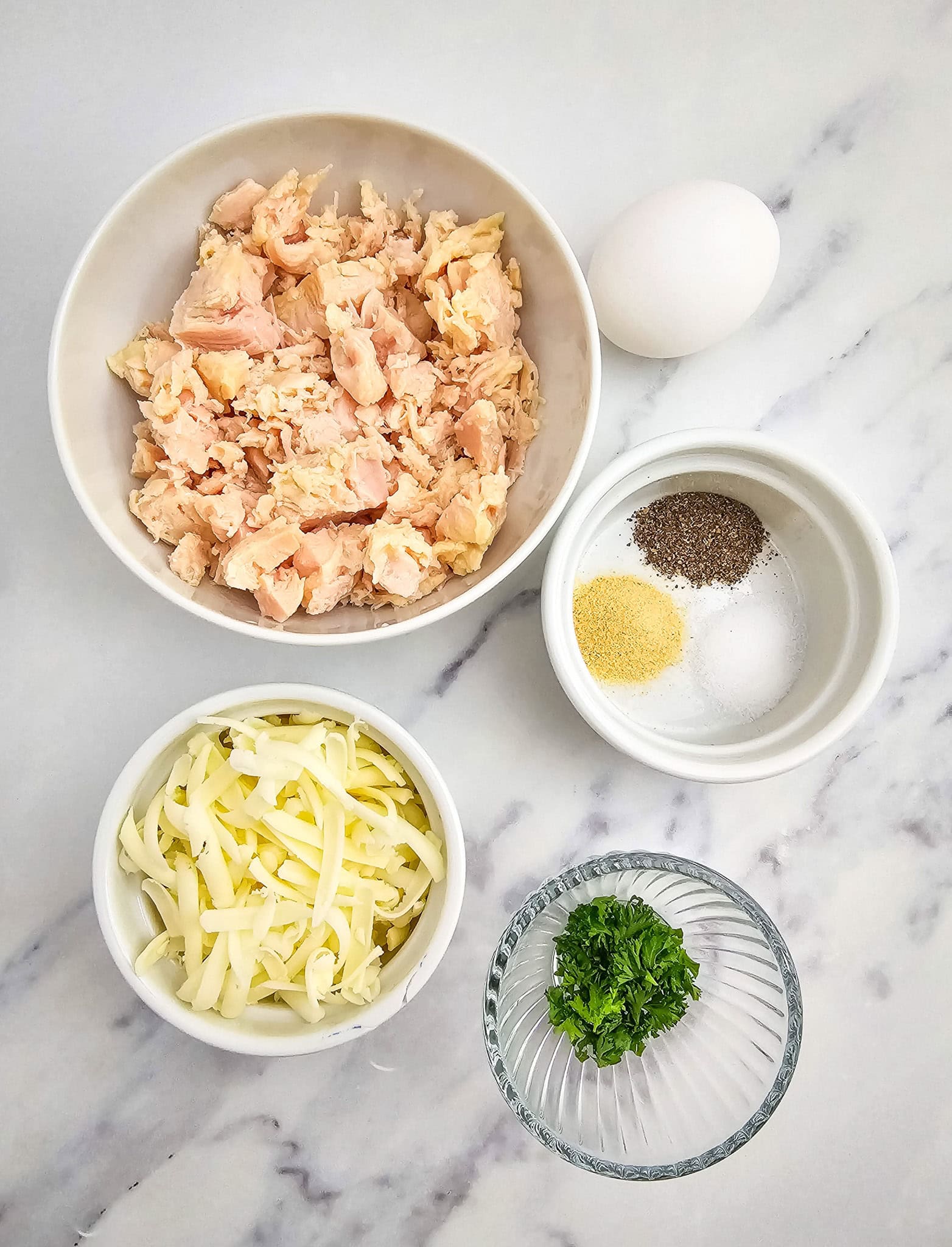 Bowls of chopped cooked chicken, shredded cheese, seasonings, chopped parsley, and a whole egg on a marble surface