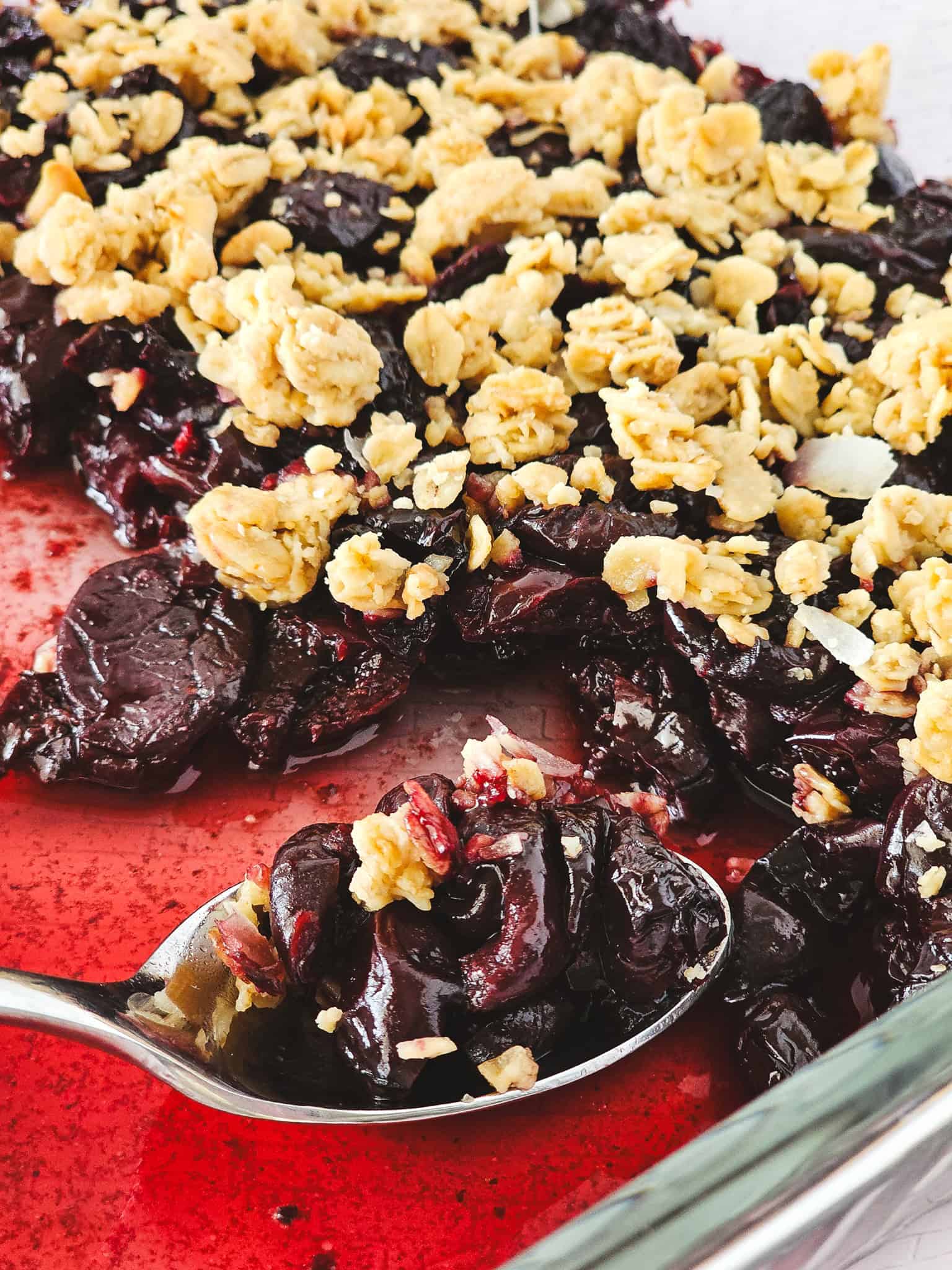 A spoon scoops cherry crisp with oats from a baking dish, showing juicy frozen cherries and crumbly topping—an effortless dessert made even easier in the air fryer.