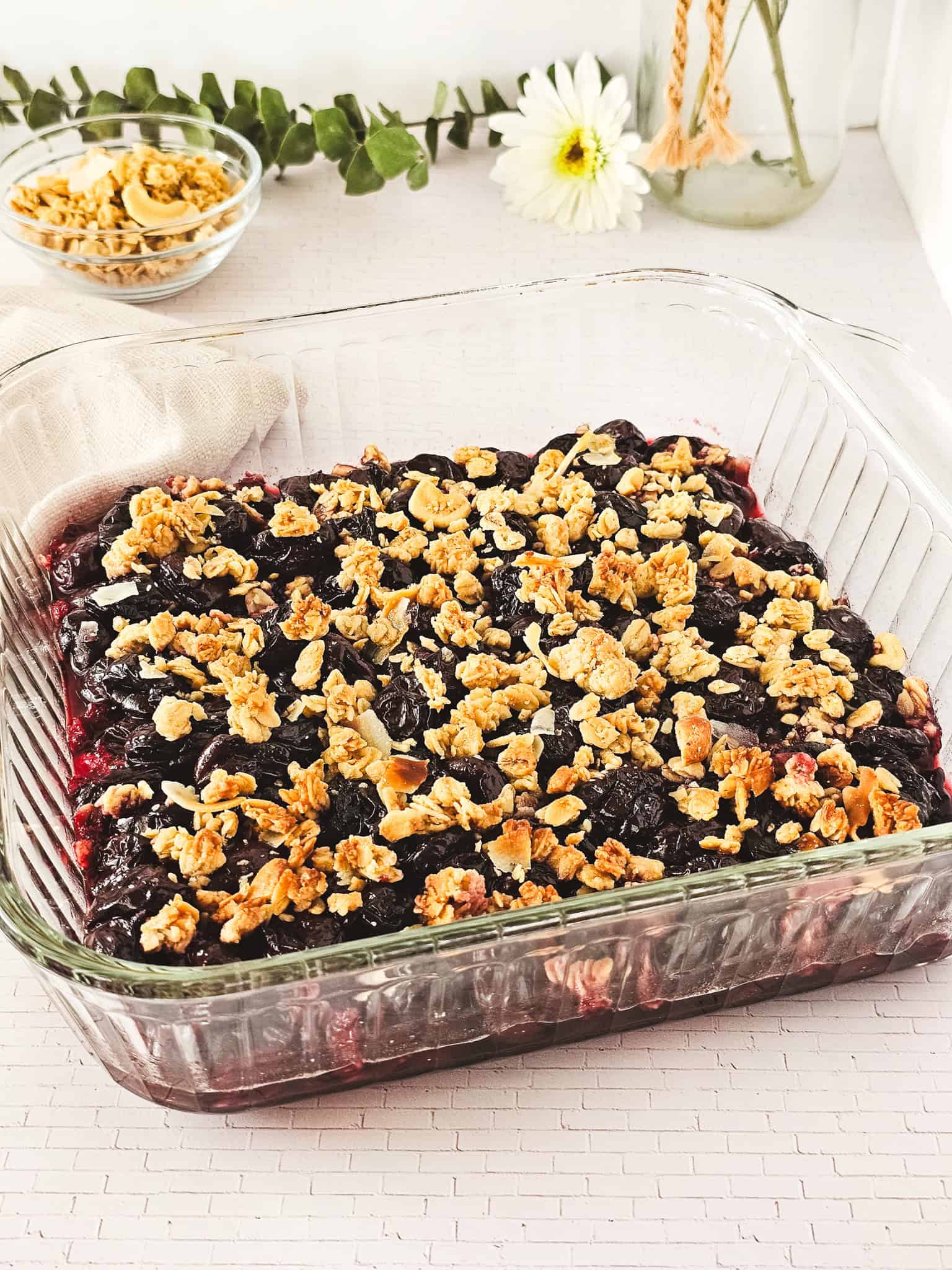 A glass dish filled with cherry crisp made from frozen cherries sits on a white table, with a flower and a bowl of granola nearby.