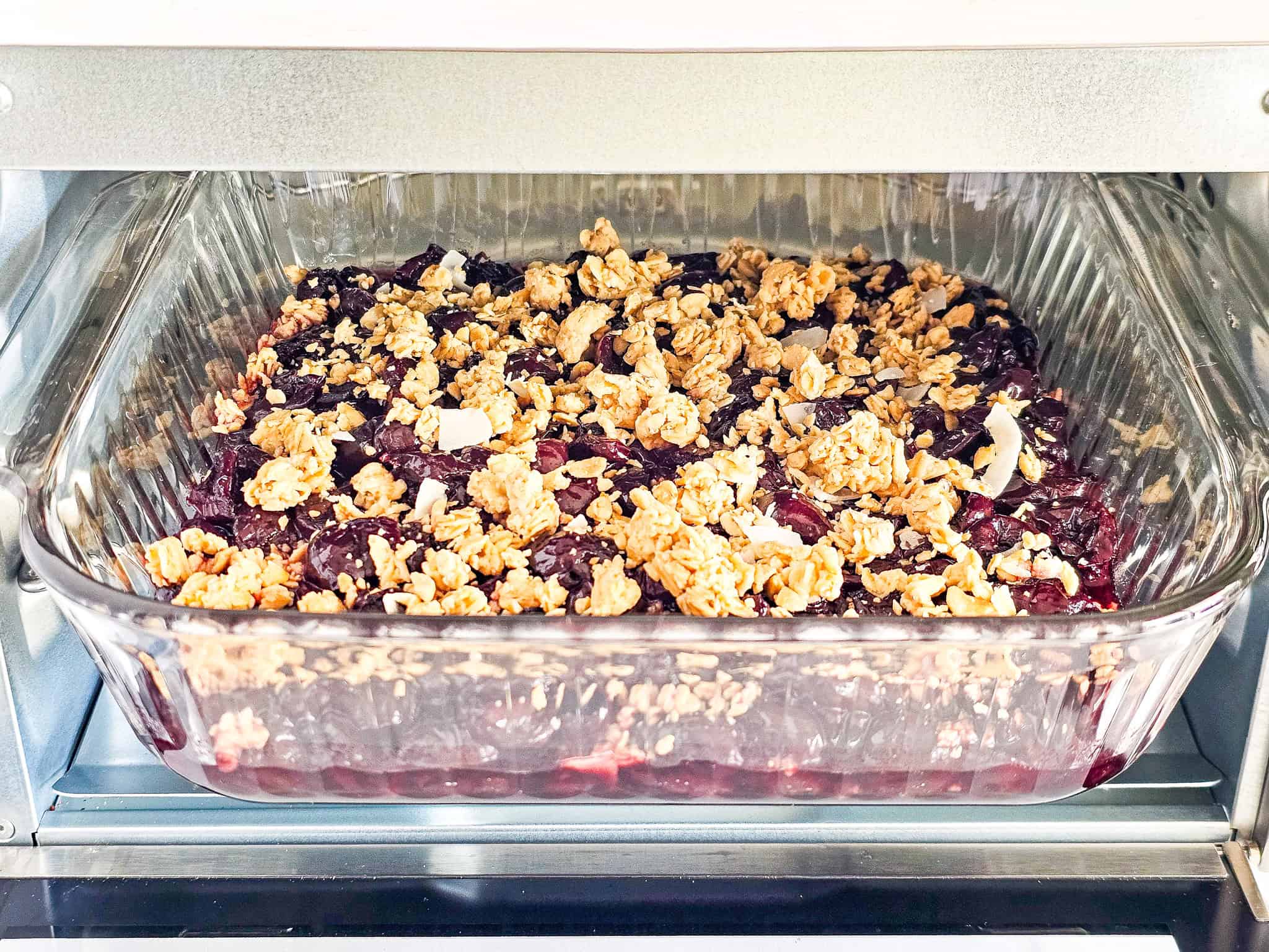 A glass baking dish with cherry crisp made from frozen cherries and topped with granola, baking in an oven.