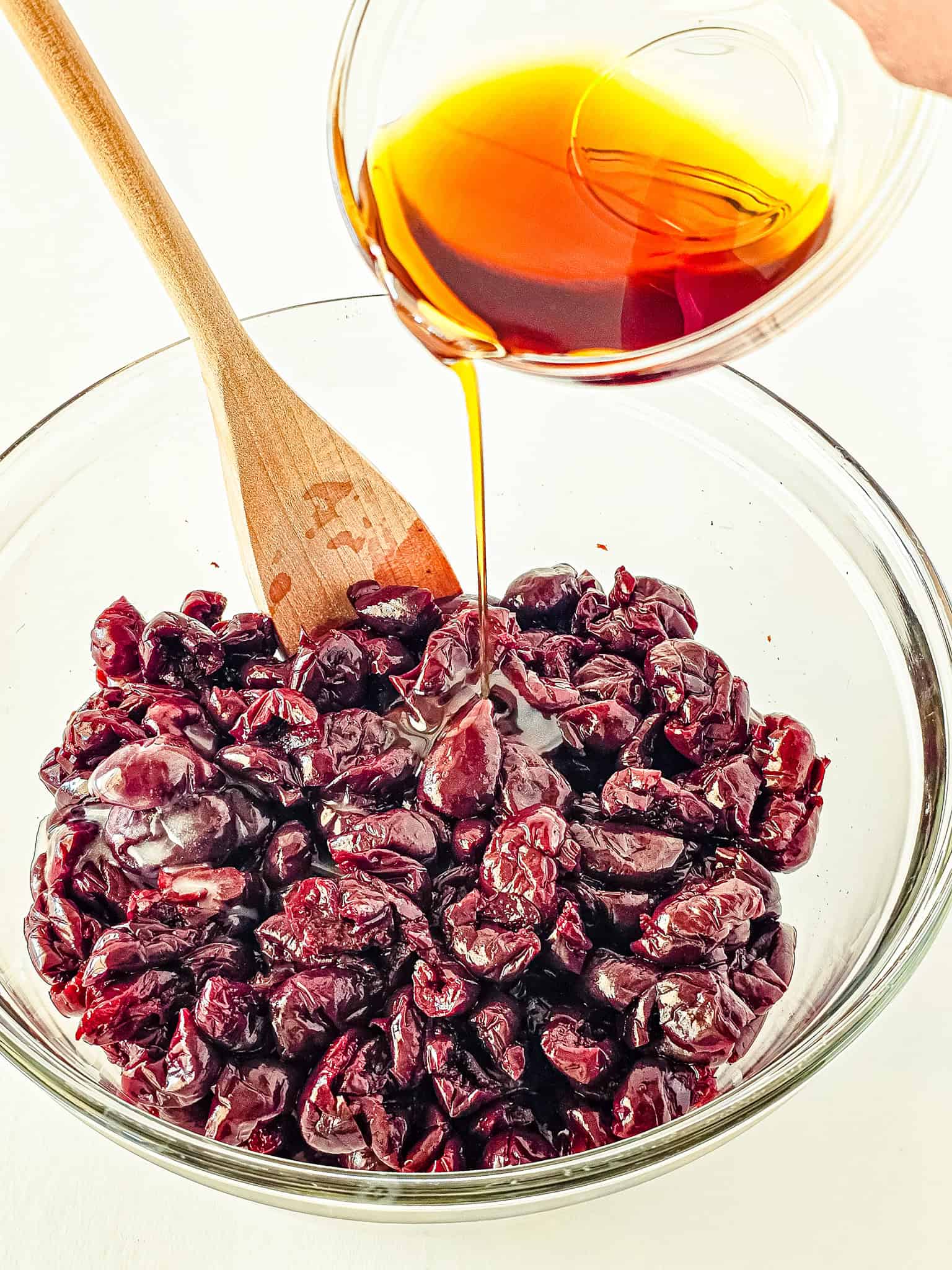 Maple syrup is poured into a glass bowl of dried cranberries and frozen cherries, then gently stirred with a wooden spoon.