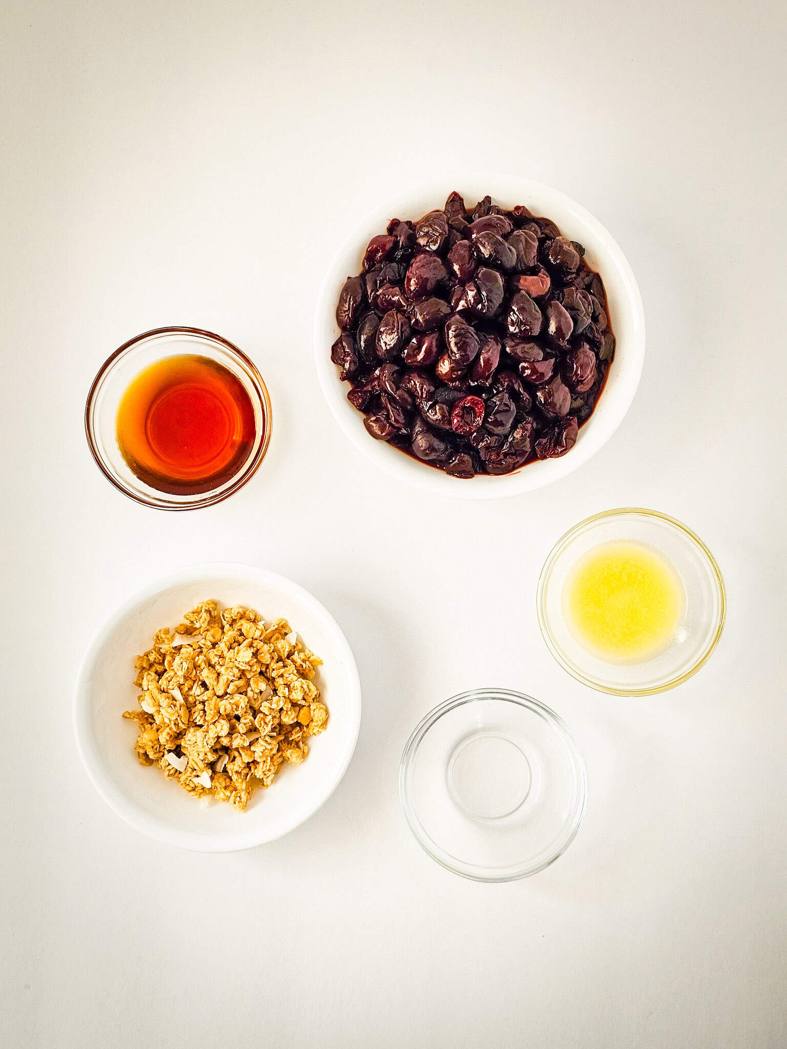 Five bowls with frozen cherries, vanilla extract, lemon juice, granola, and water on a white background—perfect ingredients for a quick cherry crisp or an easy air fryer dessert.