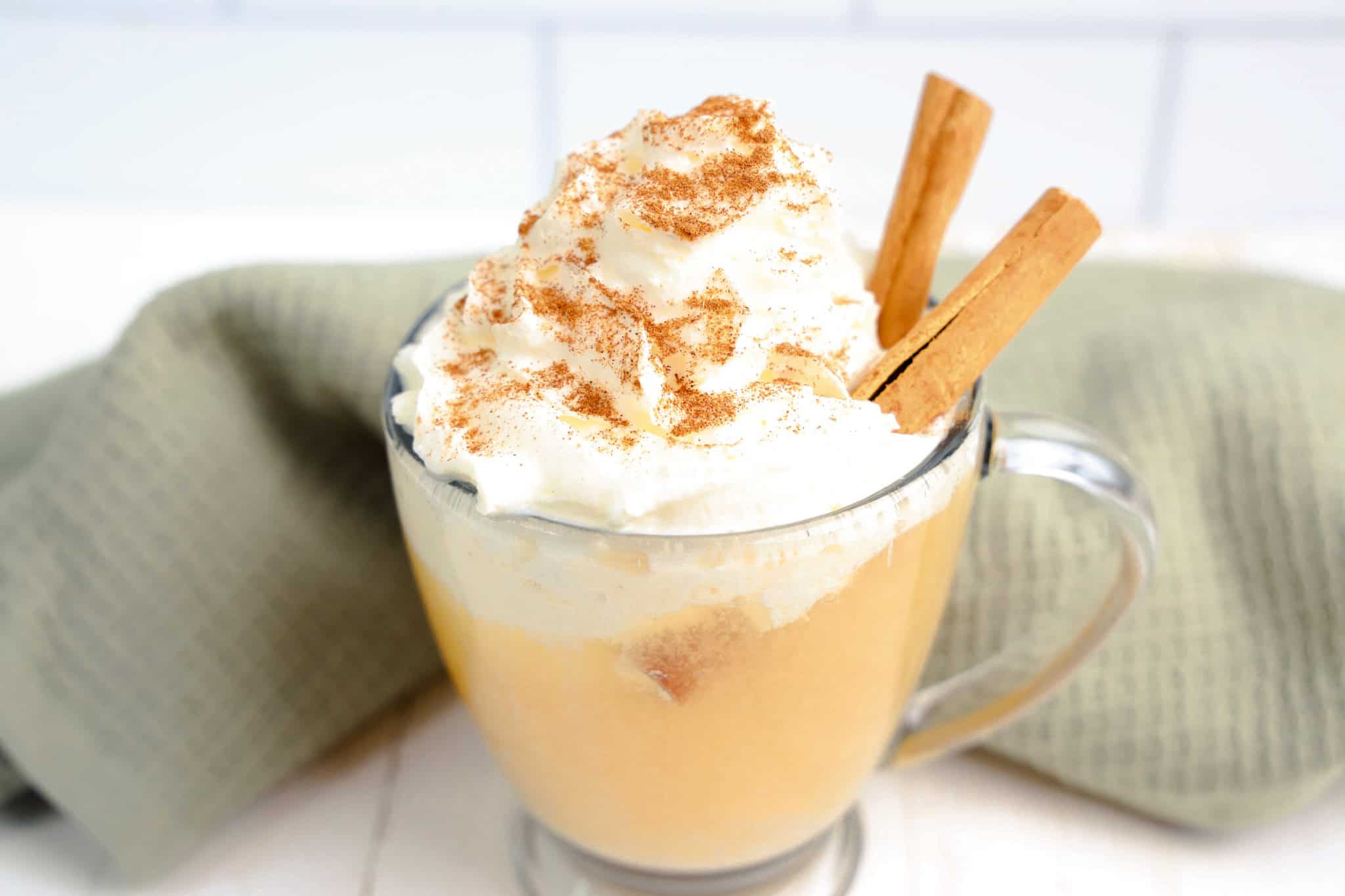 A glass mug of apple cider float with whipped cream, cinnamon, and two cinnamon sticks sits beside a green cloth