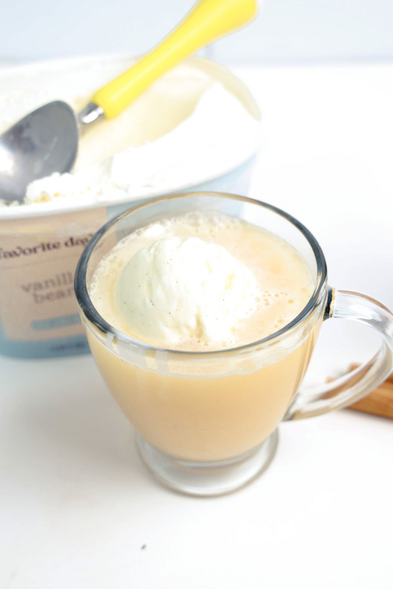 A glass mug with a scoop of vanilla ice cream and apple cider creates a tempting float, with an ice cream tub and yellow scoop in the background.