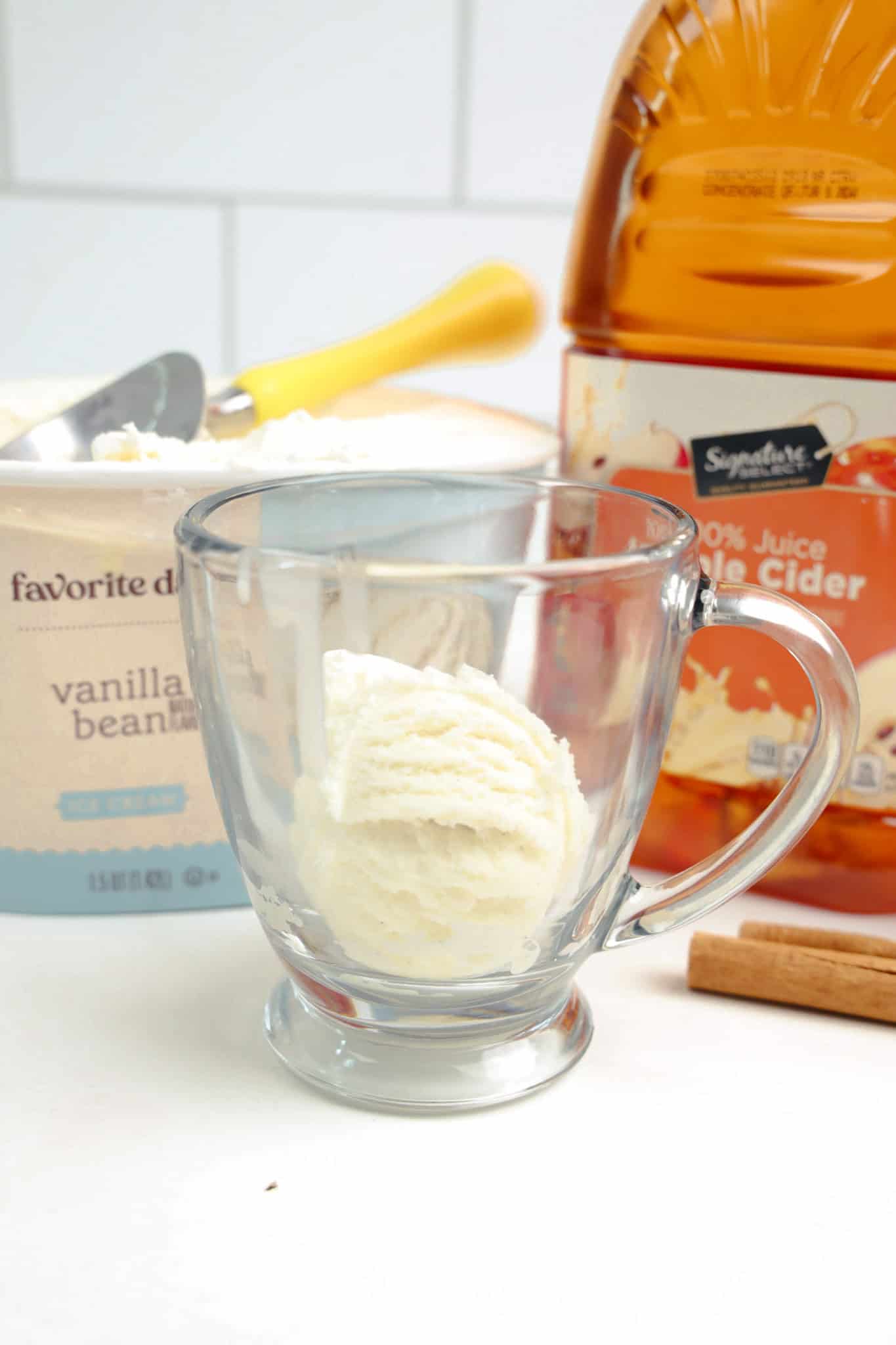 A glass mug with a scoop of vanilla ice cream, next to creamy vanilla ice cream, apple cider, and a cinnamon stick