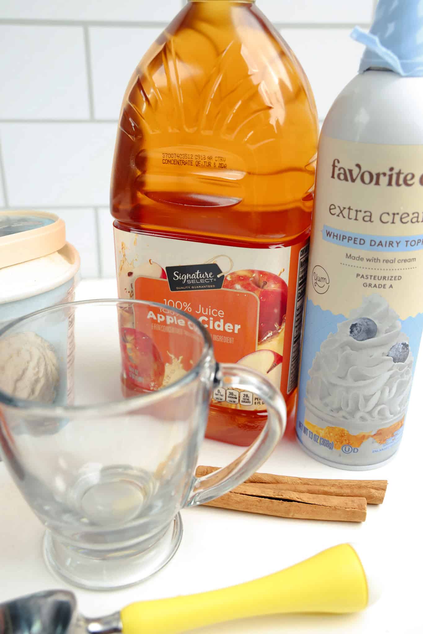 Apple Cider Float ingredients with apple cider, whipped cream, vanilla ice cream, and cinnamon sticks, served in a glass mug on a white countertop.