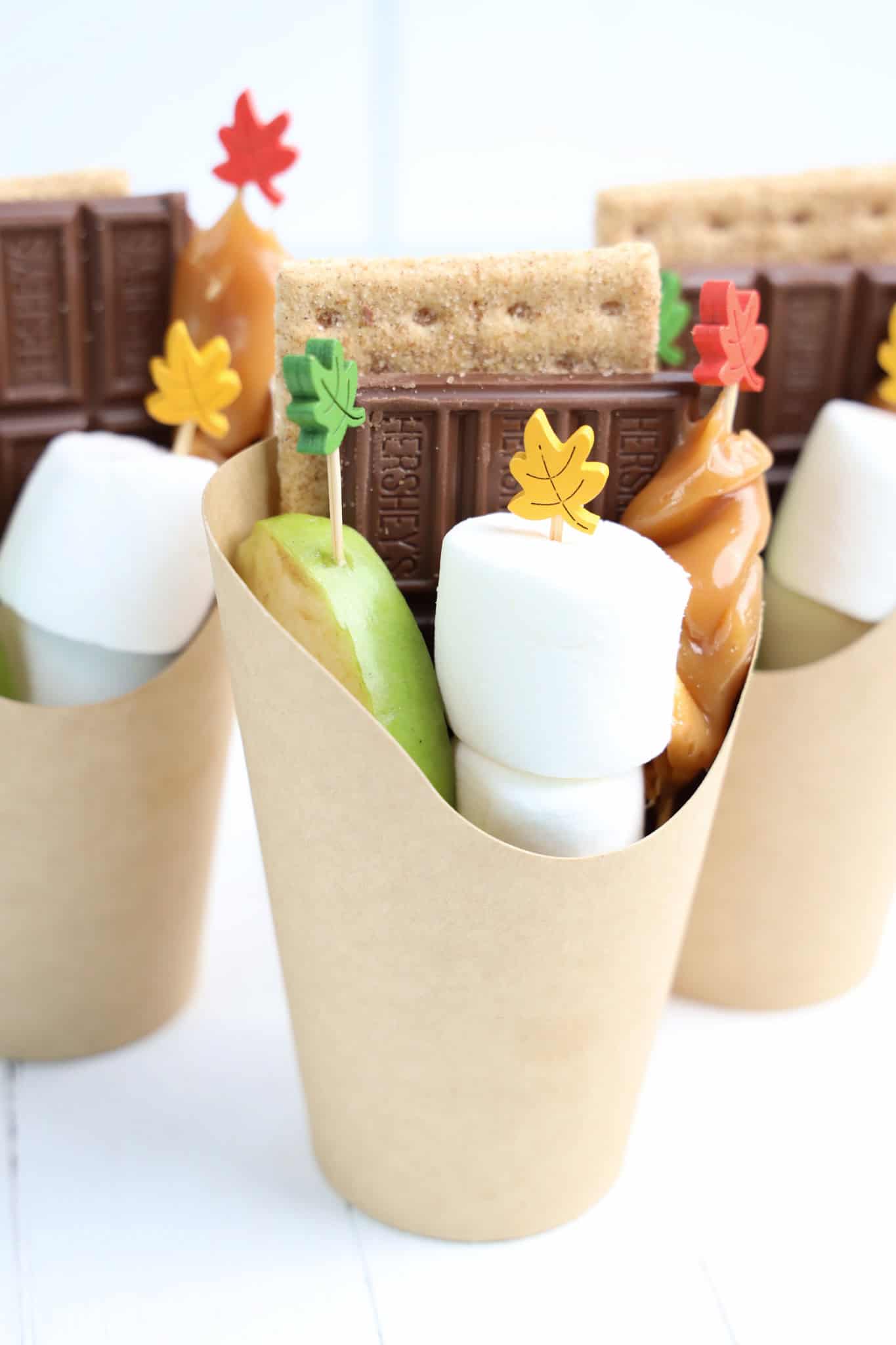 paper cups filled with apple slices, marshmallows, chocolate, and s’mores ingredients, all topped with decorative leaf picks.