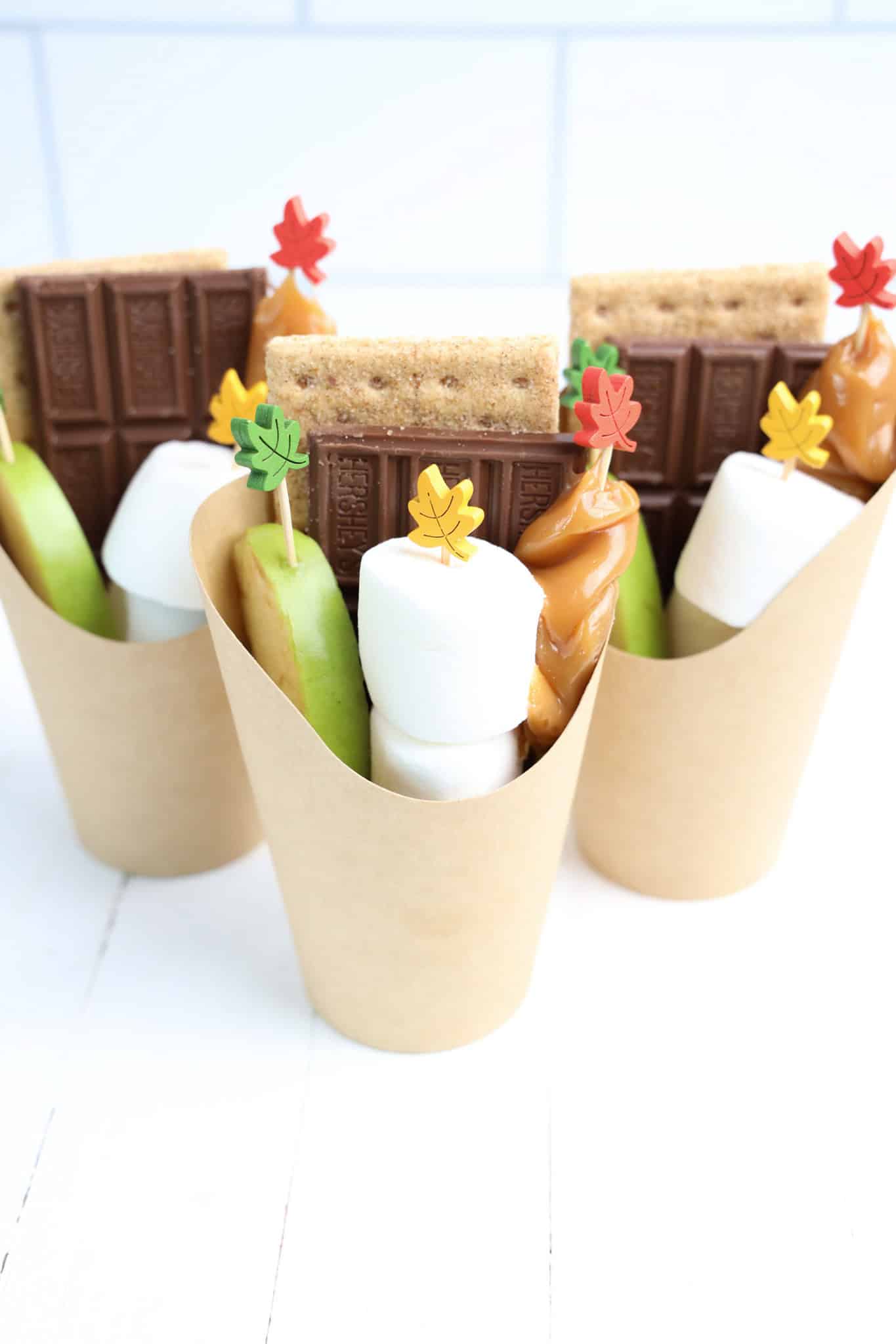 Three brown Smores Cups filled with chocolate, graham crackers, marshmallows, apple slices, and caramel each topped with colorful leaf picks.