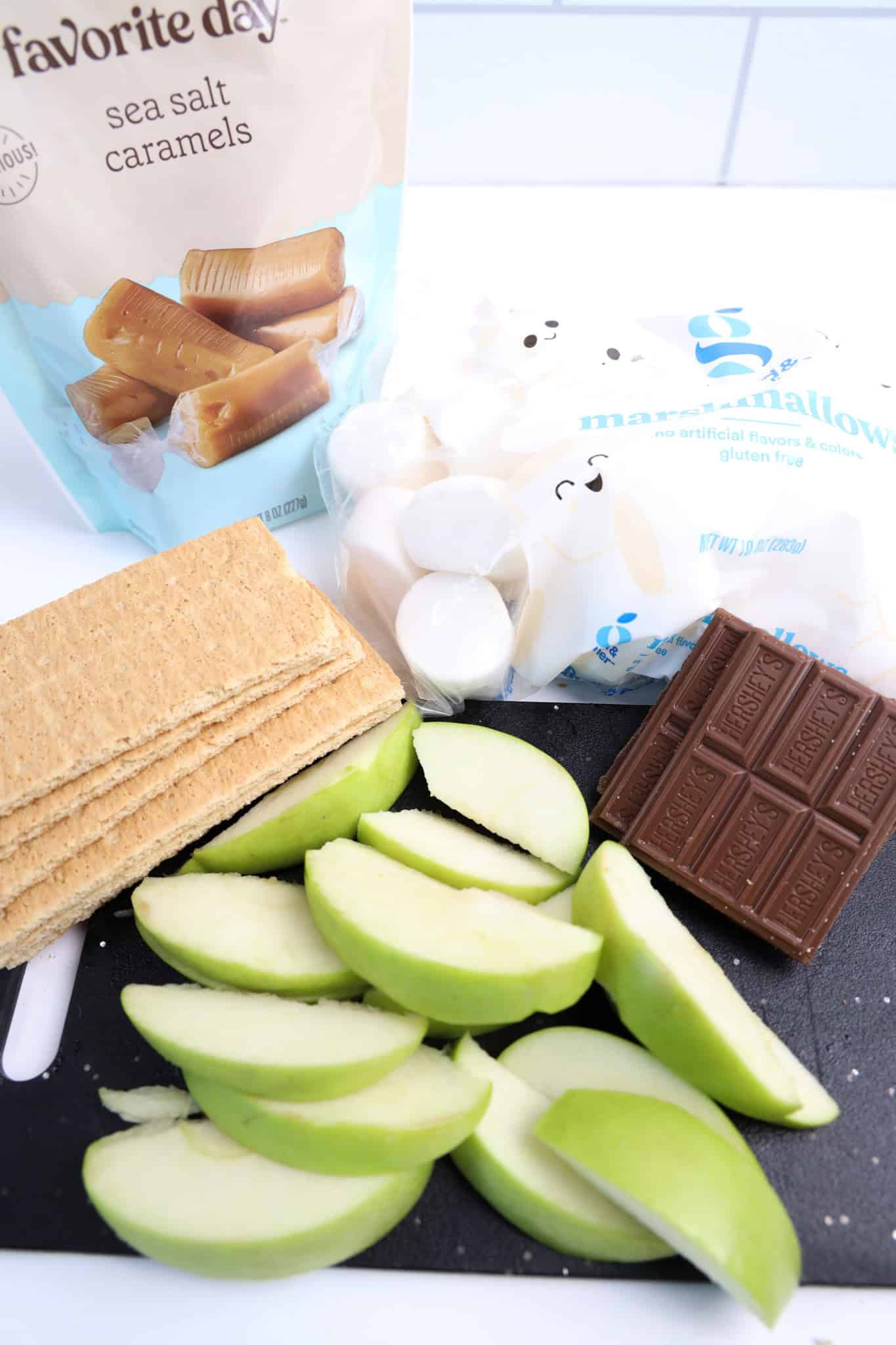 Sliced green apples, graham crackers, chocolate, caramels, and marshmallows on a cutting board