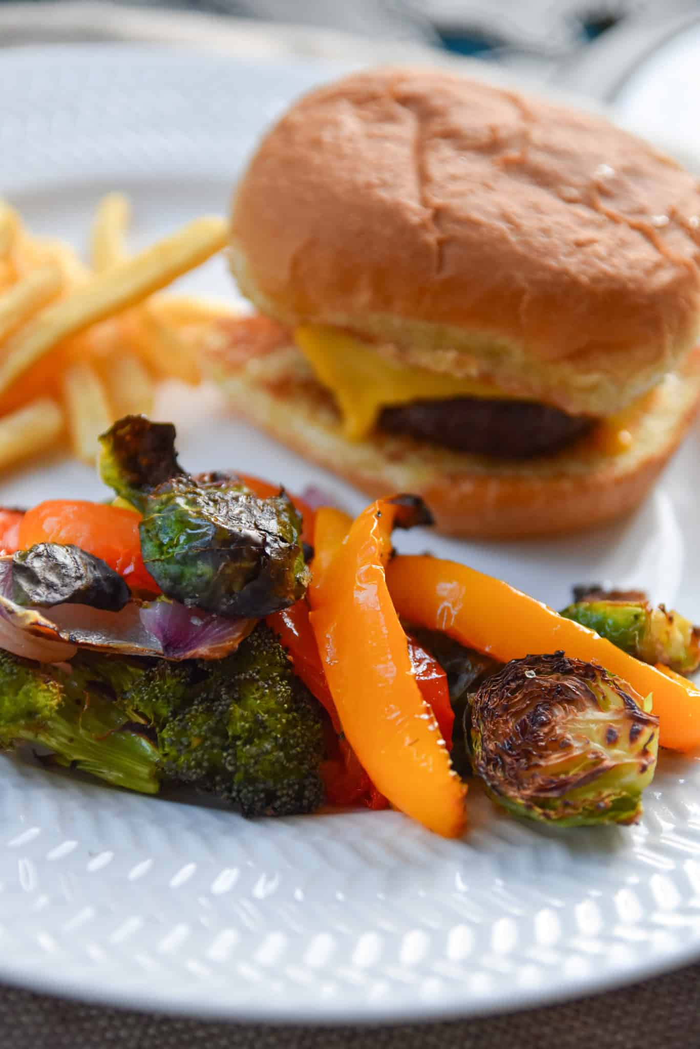 Cheeseburger with fries and a colorful mix of sheet pan roasted vegetables, including broccoli, Brussels sprouts, and bell peppers, served on a plate.