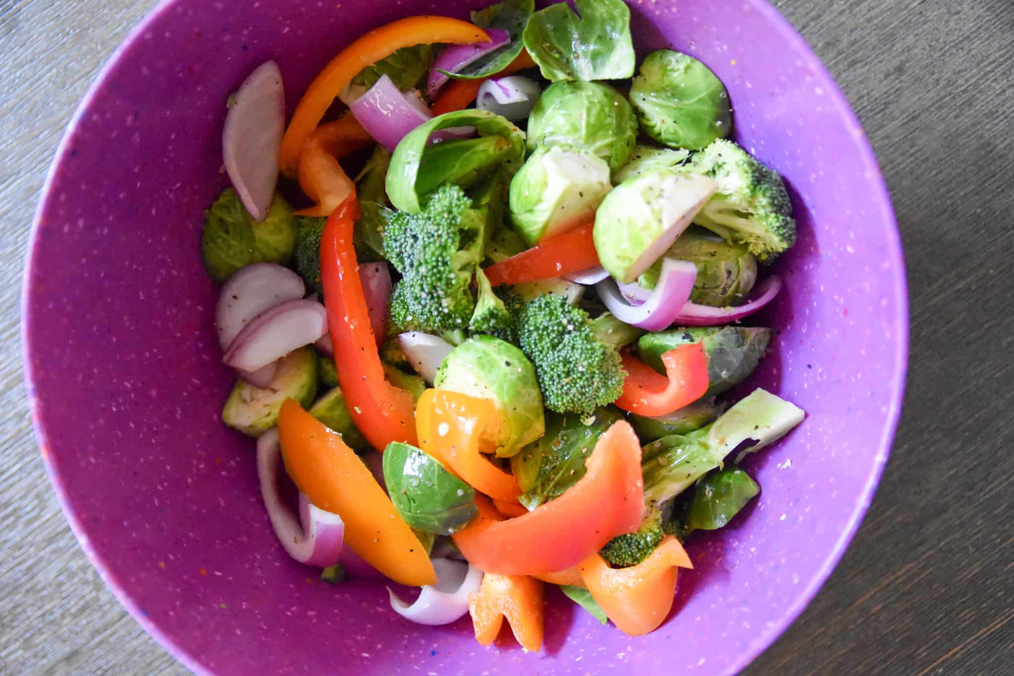 A purple bowl filled with sheet pan roasted vegetables like sliced bell peppers, broccoli, Brussels sprouts, and red onions.
