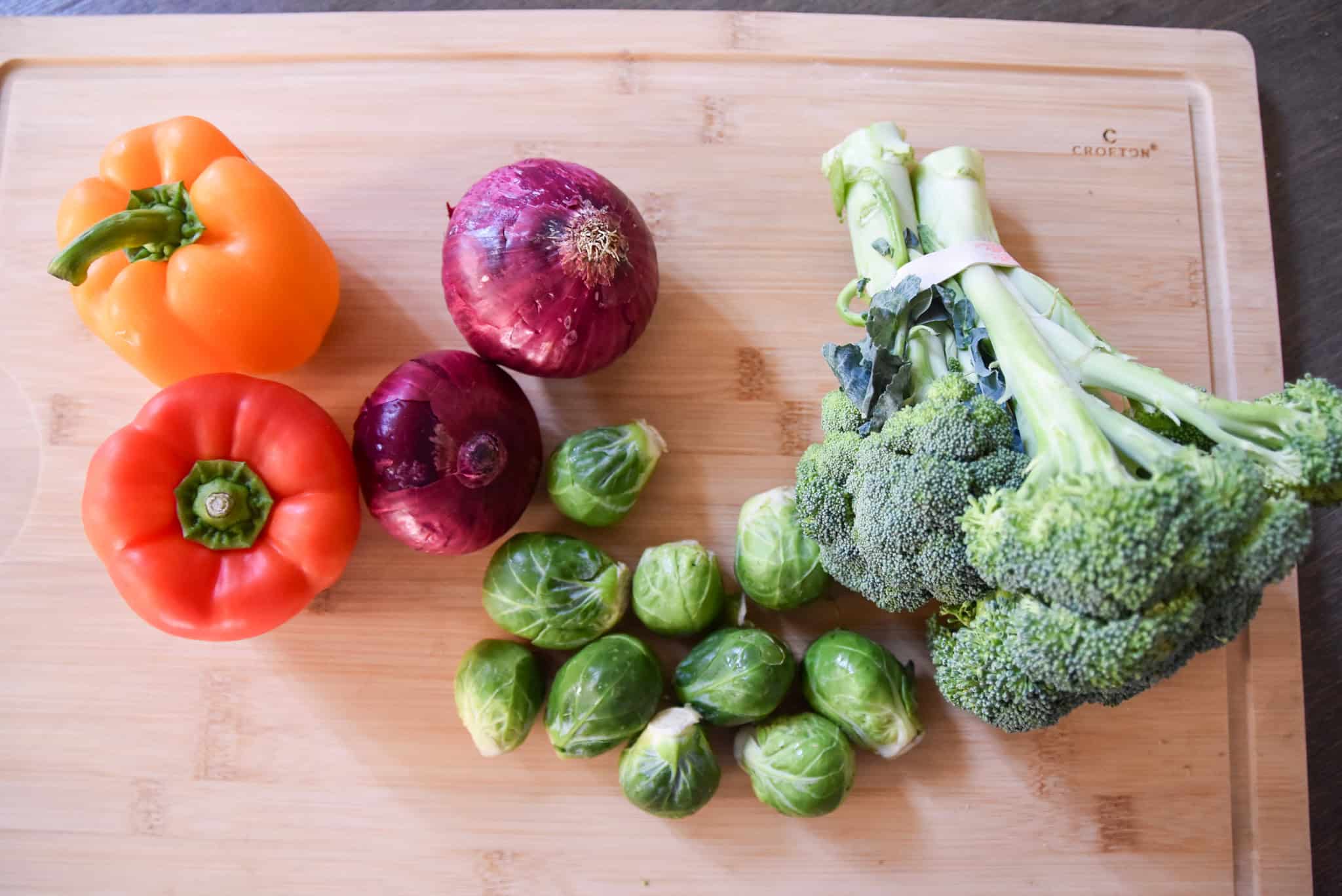 Orange and red bell peppers, two red onions, brussels sprouts, and broccoli are perfect for sheet pan roasted vegetables on a wooden cutting board.