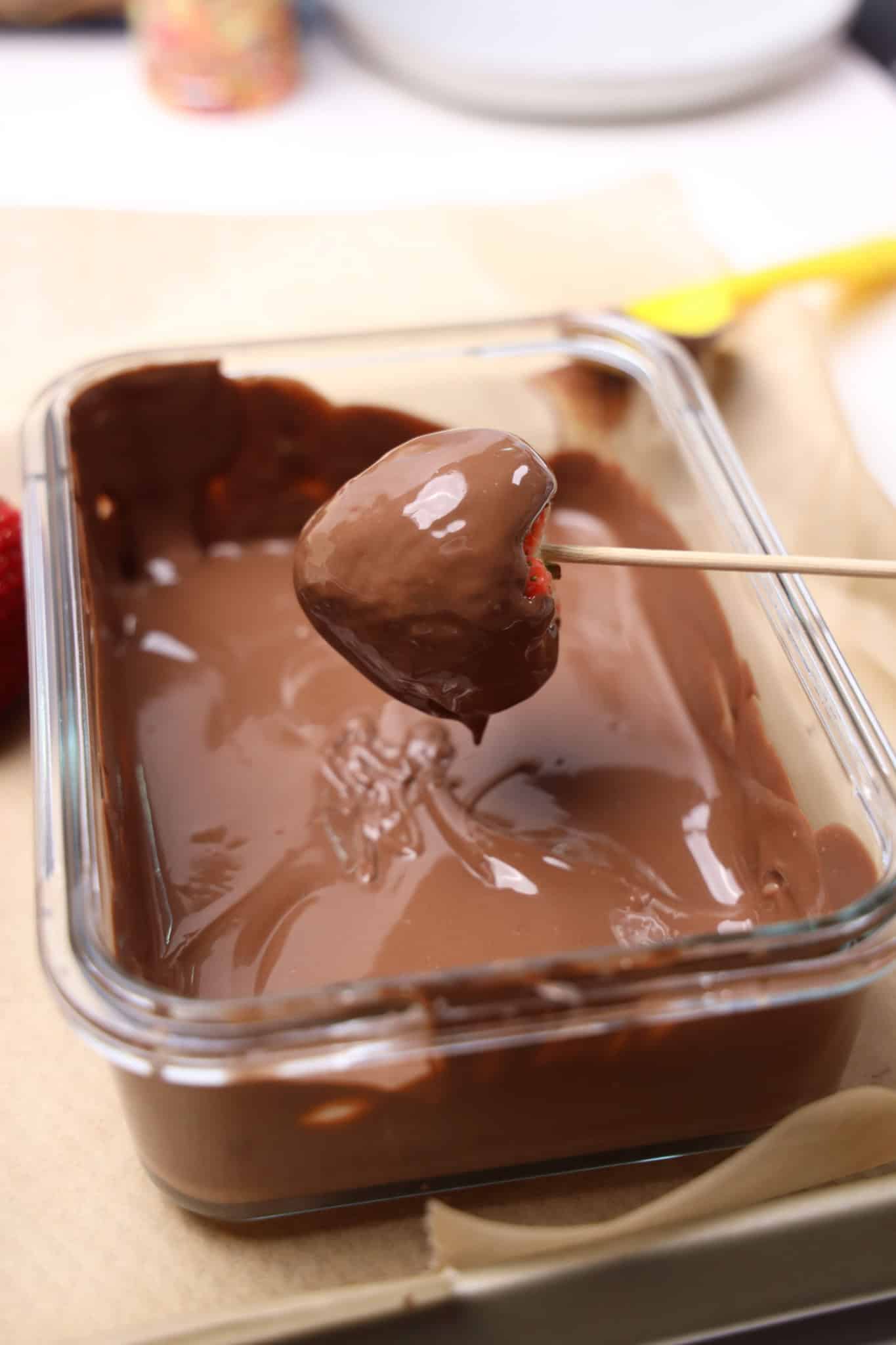 A strawberry on a stick is dipped into melted chocolate in a glass container, creating delicious chocolate covered strawberries.