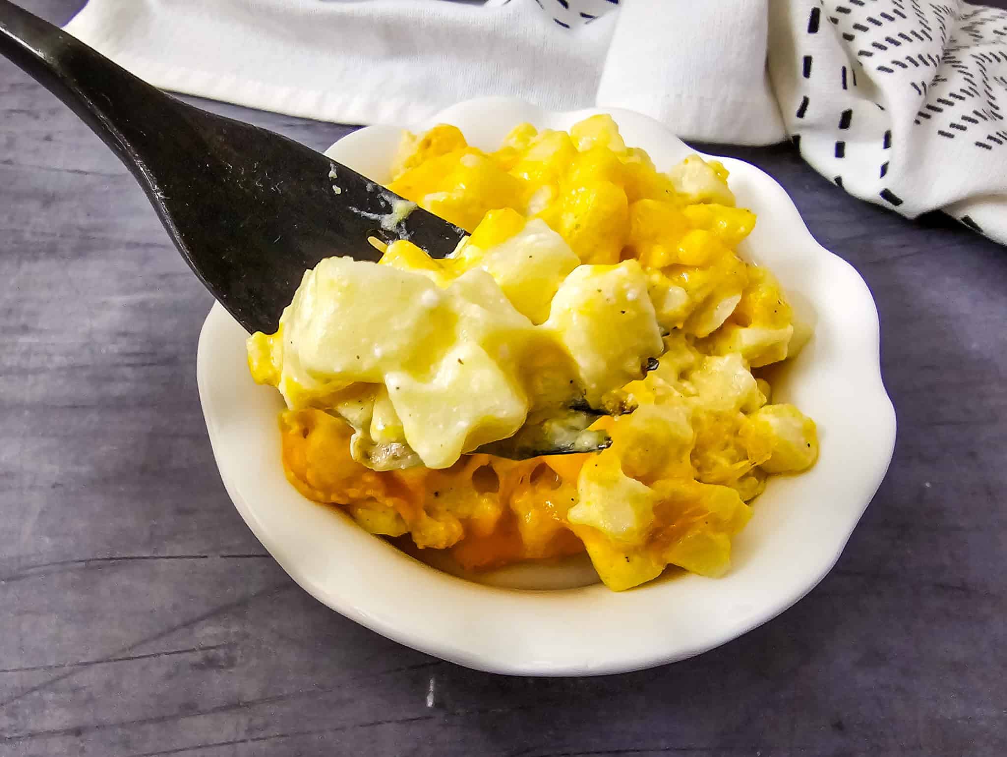 A fork lifts a serving of cheesy baked macaroni, cauliflower, and potatoes from a white bowl on a gray table.