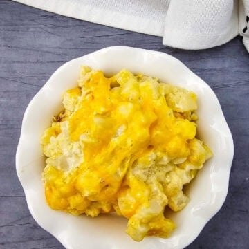 A white bowl filled with Crockpot cheesy potatoes sits on a gray surface beside a white patterned cloth.