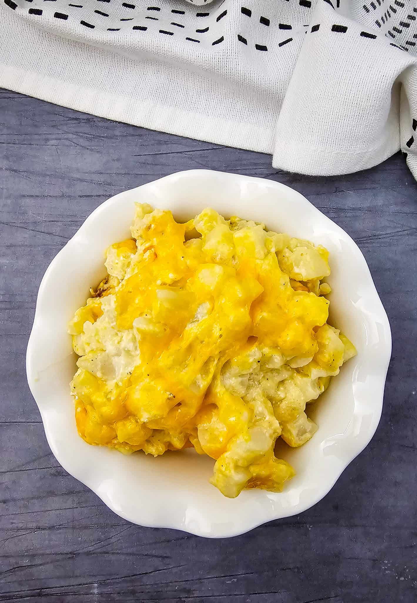A white bowl filled with Crockpot cheesy potatoes sits on a gray surface beside a white patterned cloth.