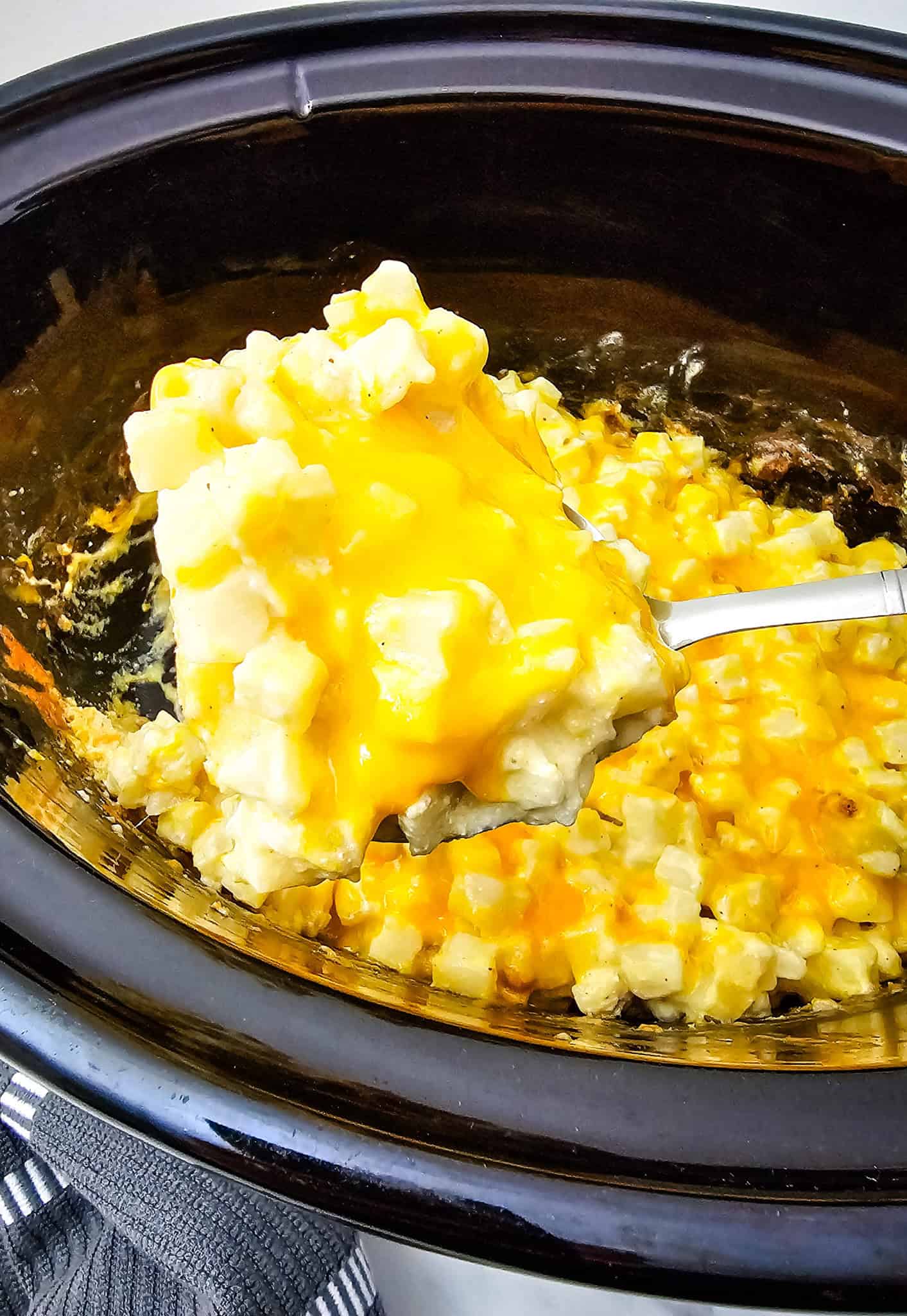 A spoonful of cheesy potatoes casserole being served from a black Crockpot.