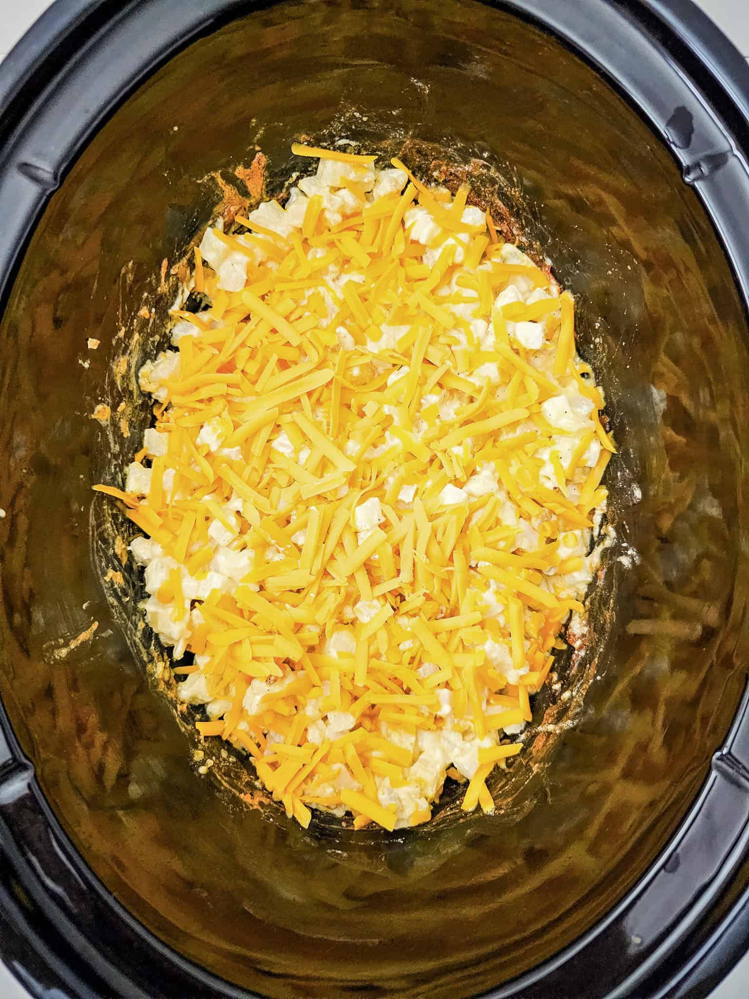 Shredded cheddar cheese tops a creamy mixture of Crockpot Cheesy Potatoes, all inside a black slow cooker.