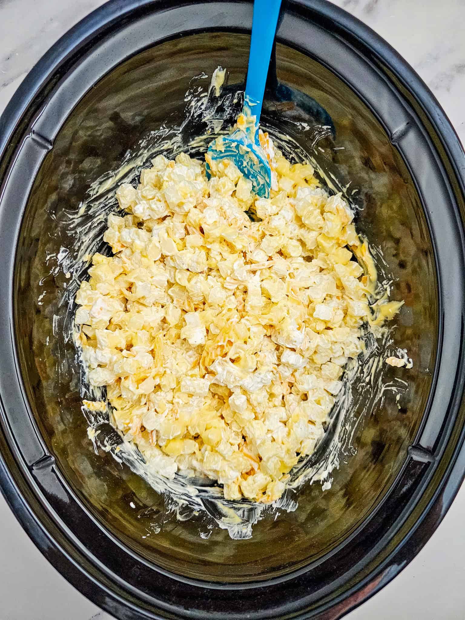 Creamy Crockpot Cheesy Potatoes with diced potatoes in a rich sauce, shown in a black slow cooker and stirred with a blue spatula.