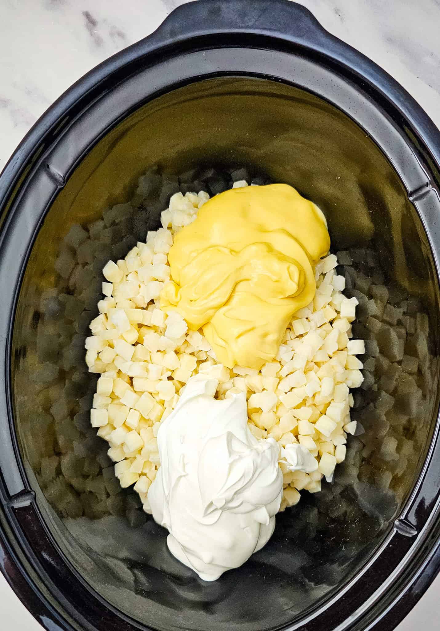 Diced potatoes, sour cream, and cream soup in a black crockpot on a marble countertop-perfect for making Crockpot Cheesy Potatoes.