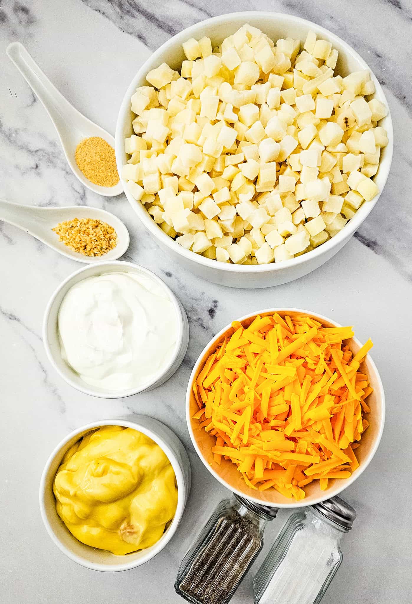 Bowls of diced potatoes, shredded cheese, sour cream, soup, and spices are ready on a marble countertop for easy Slow Cooker Cheesy Potatoes.