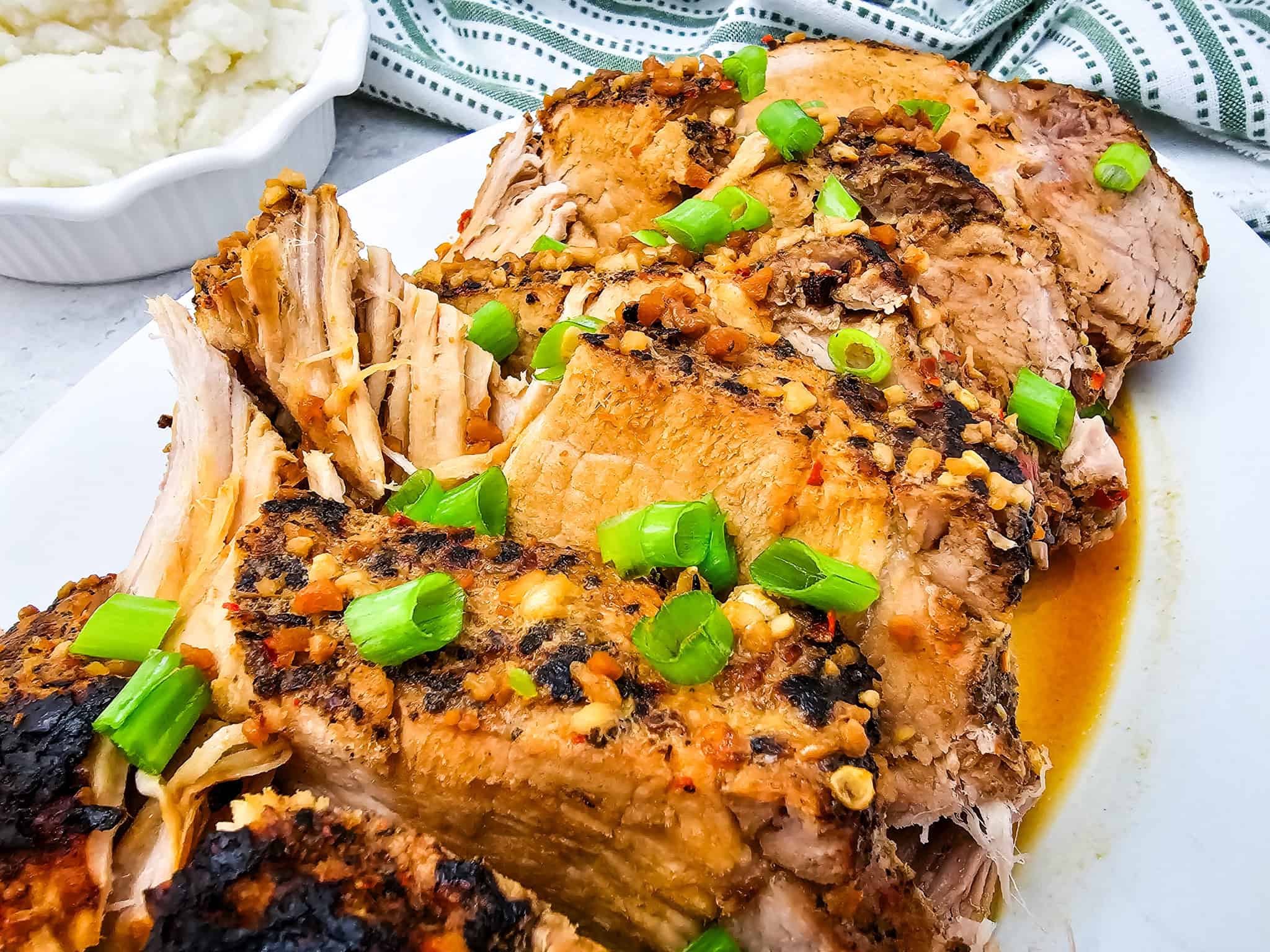 Sliced slow cooker pork topped with chopped green onions on a white plate, with mashed potatoes in the background.