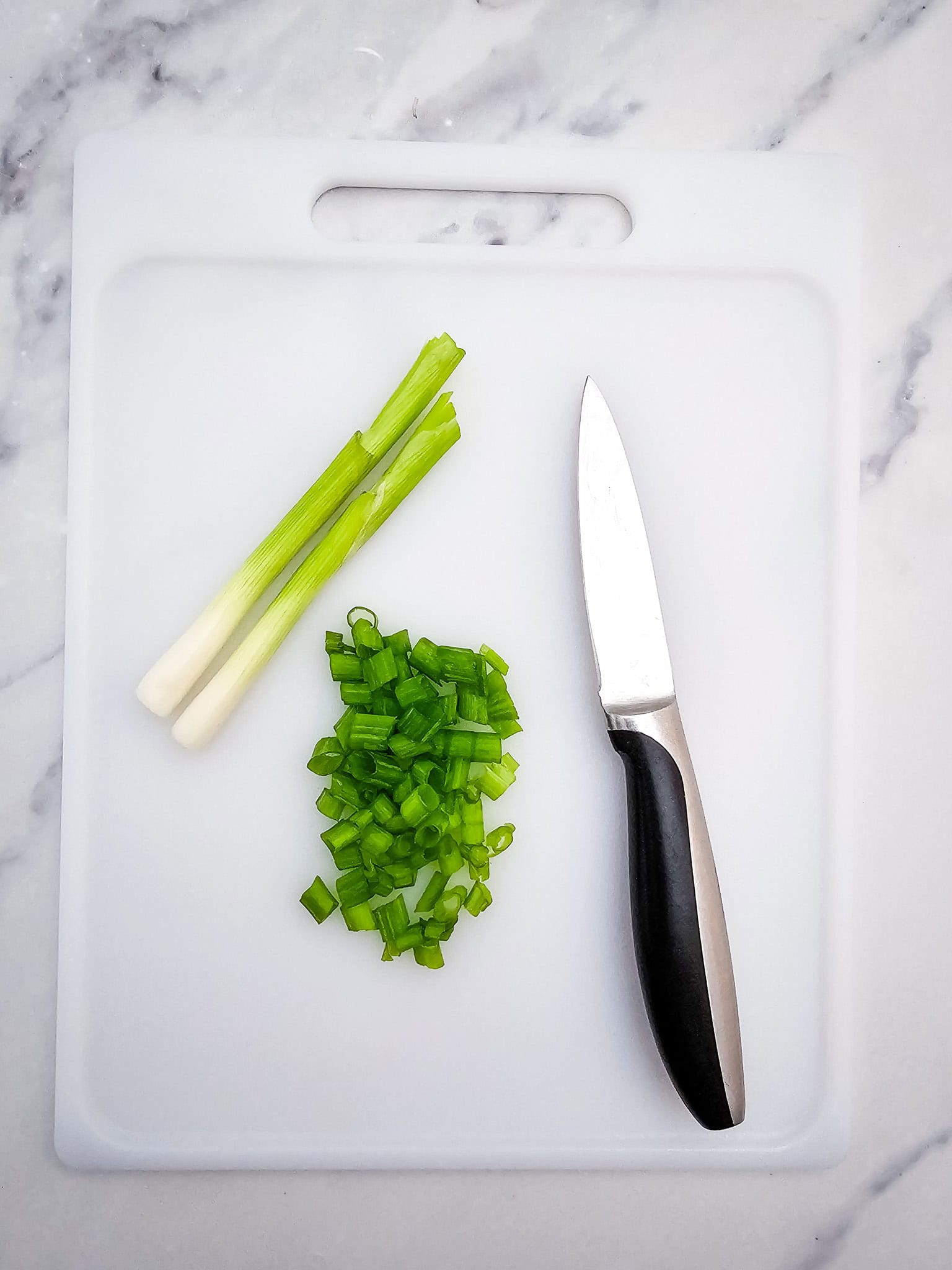 Chopped green onions, whole scallions, and a knife on a white cutting board-perfect garnishes for your Honey Garlic Pork Roast cooked in the crockpot, all set against a marble surface.