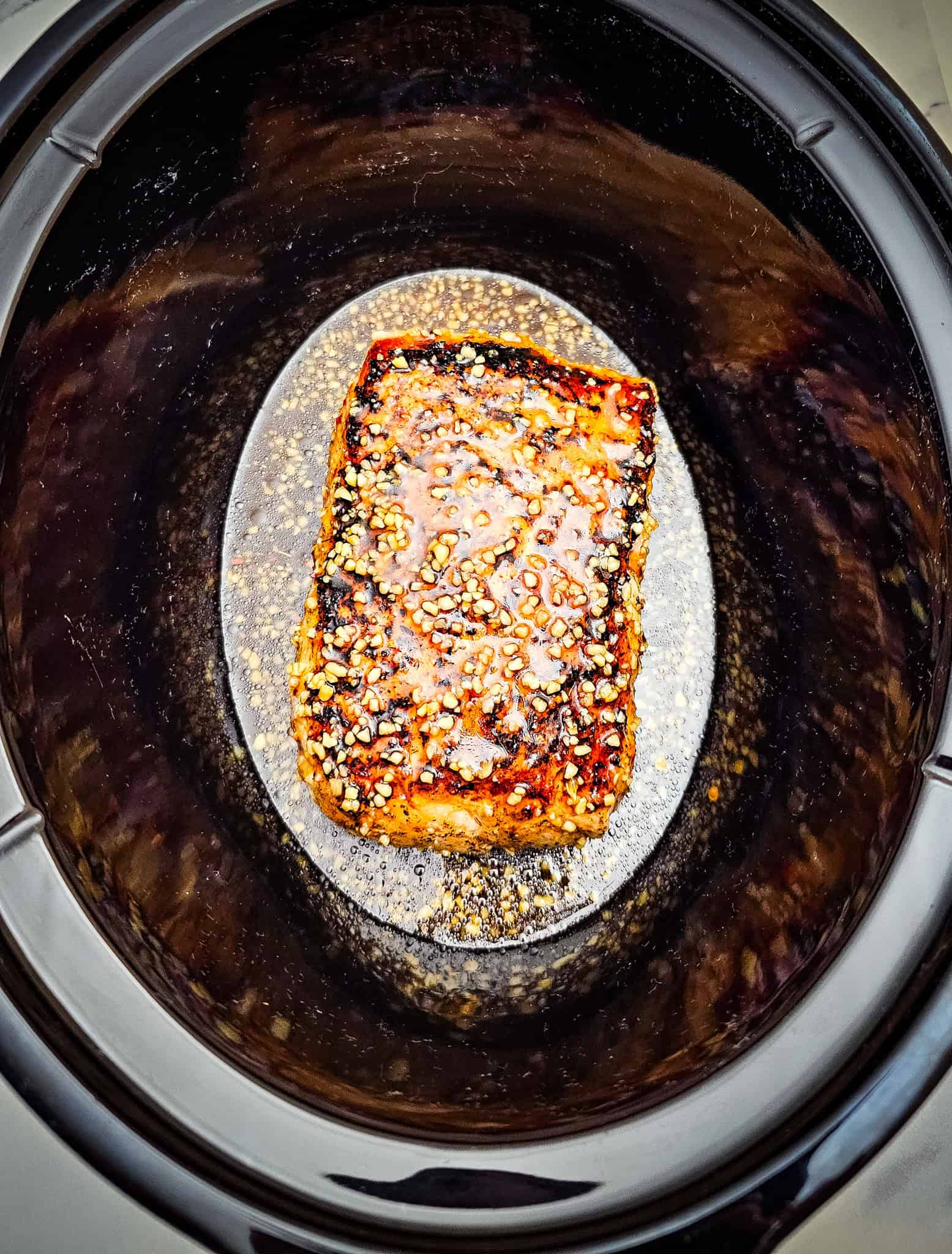 A glazed piece of salmon with sesame seeds sits in the center of a black Crockpot, ready to impress.