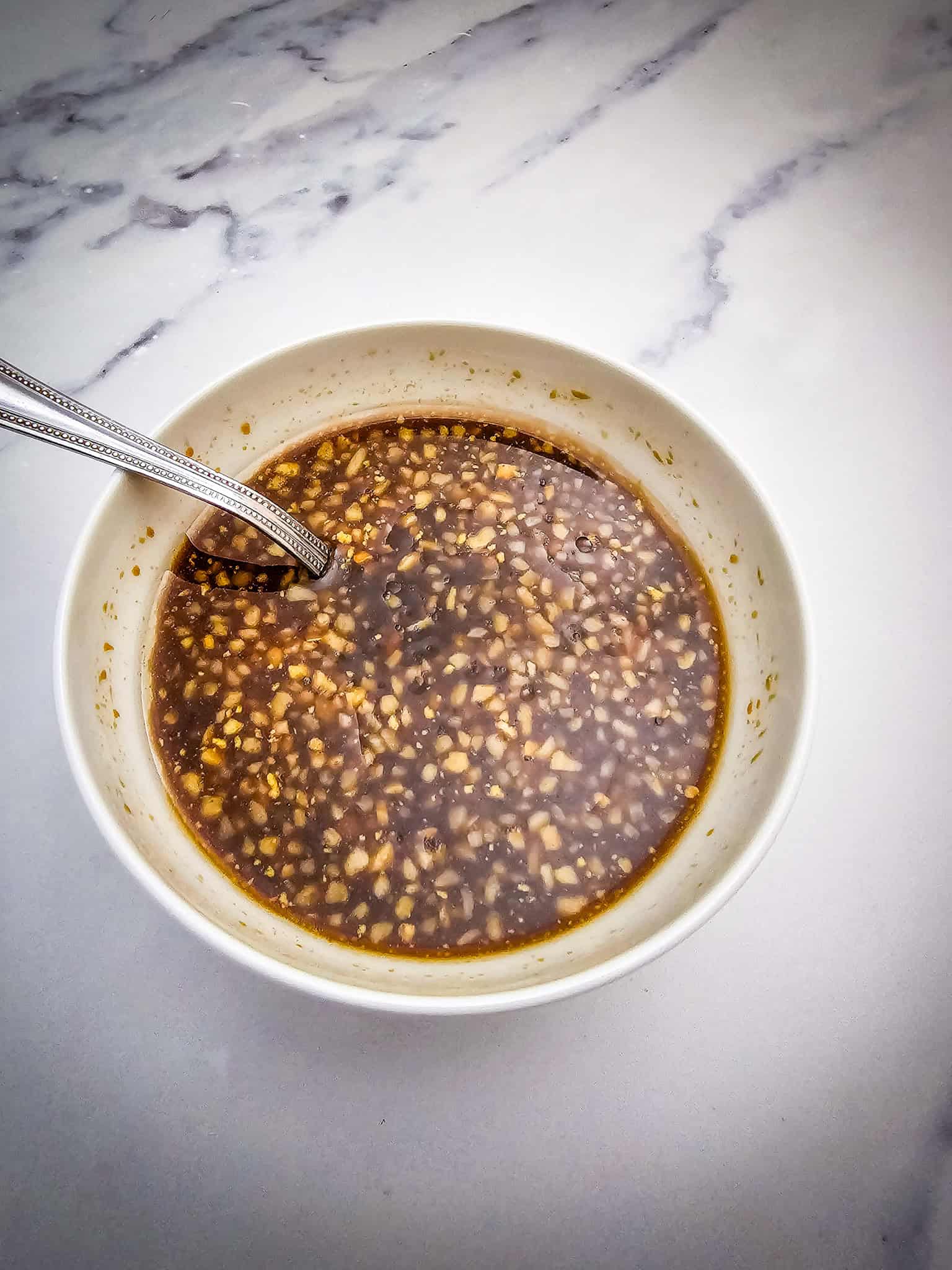 A bowl of brown, chunky sauce from a Crockpot Honey Garlic Pork Roast sits with a spoon on a white marble surface.