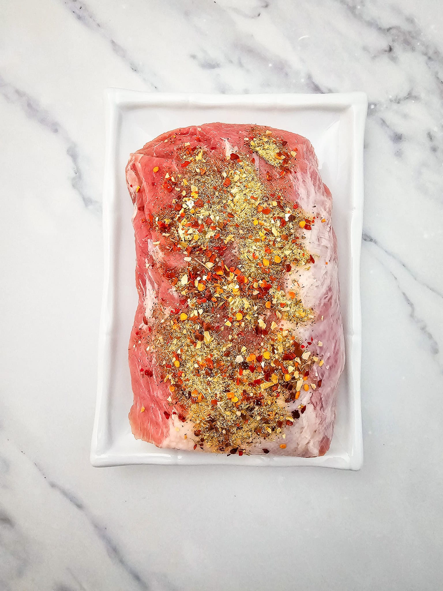 Raw seasoned pork tenderloin on a white rectangular plate, perfect for a Honey Garlic Pork Roast in the crockpot, placed on a marble surface.