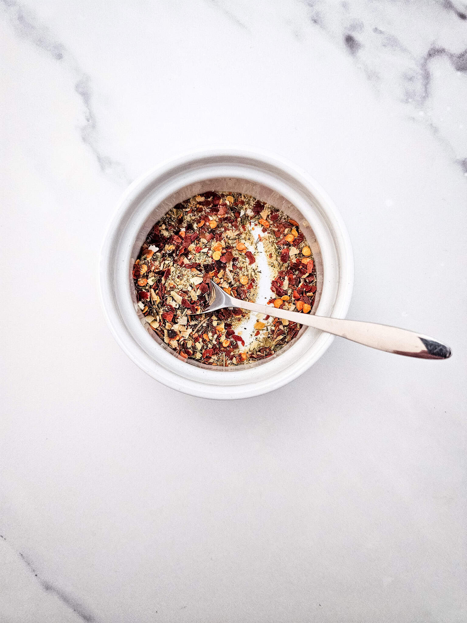 A spoon in a white bowl filled with mixed dried herbs and spices-perfect for seasoning a Honey Garlic Pork roast-sits on a white marble surface.