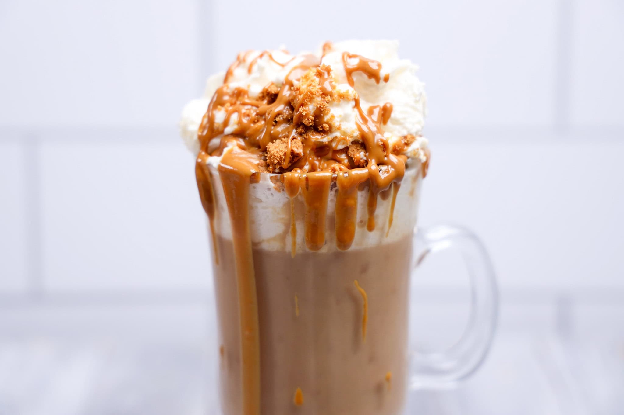 A glass mug of hot chocolate topped with whipped cream, caramel sauce, and crunchy Biscoff crumbles