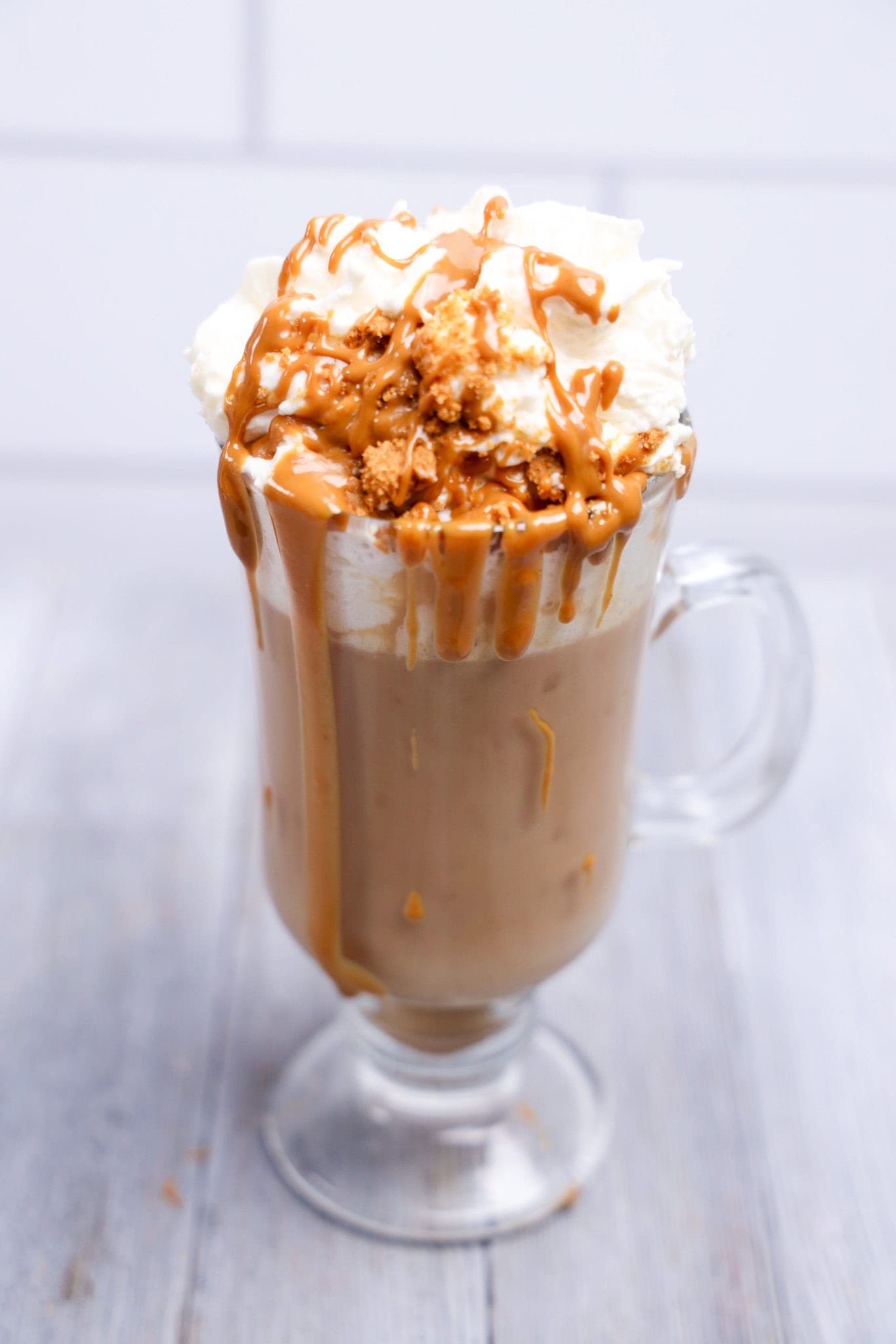 A glass mug of Biscoff Hot Chocolate topped with whipped cream, caramel drizzle, and crumbled cookie sits on a light wooden surface.