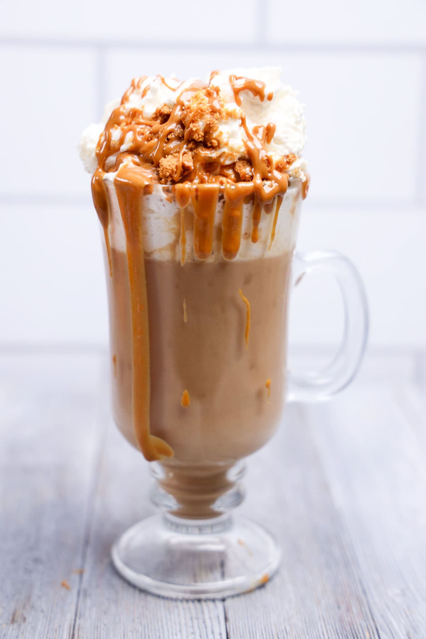 Glass mug of creamy Biscoff Hot Chocolate topped with whipped cream, caramel sauce, and crumbled cookie on a wooden table.